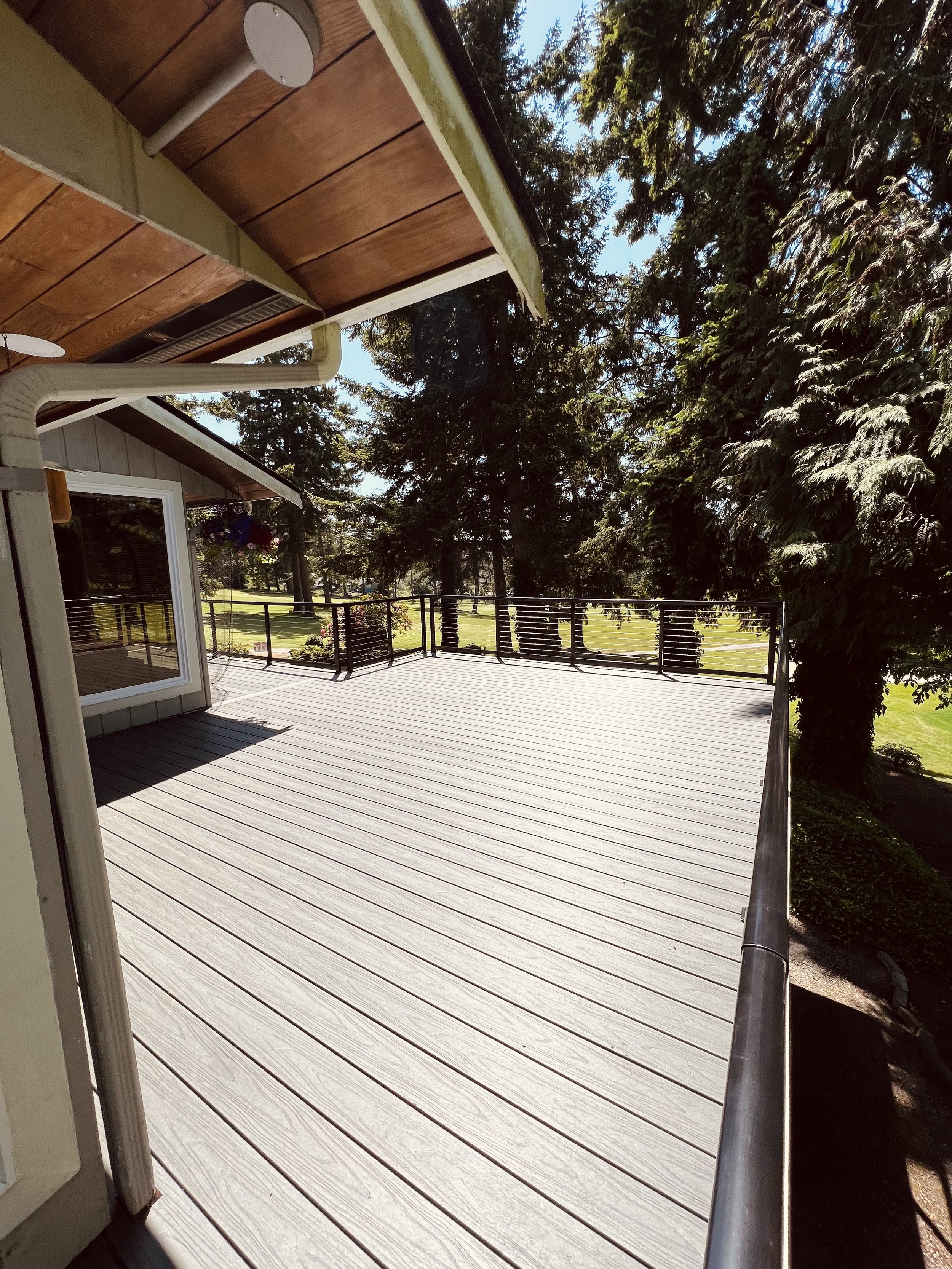 Empty wooden backyard deck with black metal railing, surrounded by tall green trees and a grassy yard in bright sunlight.