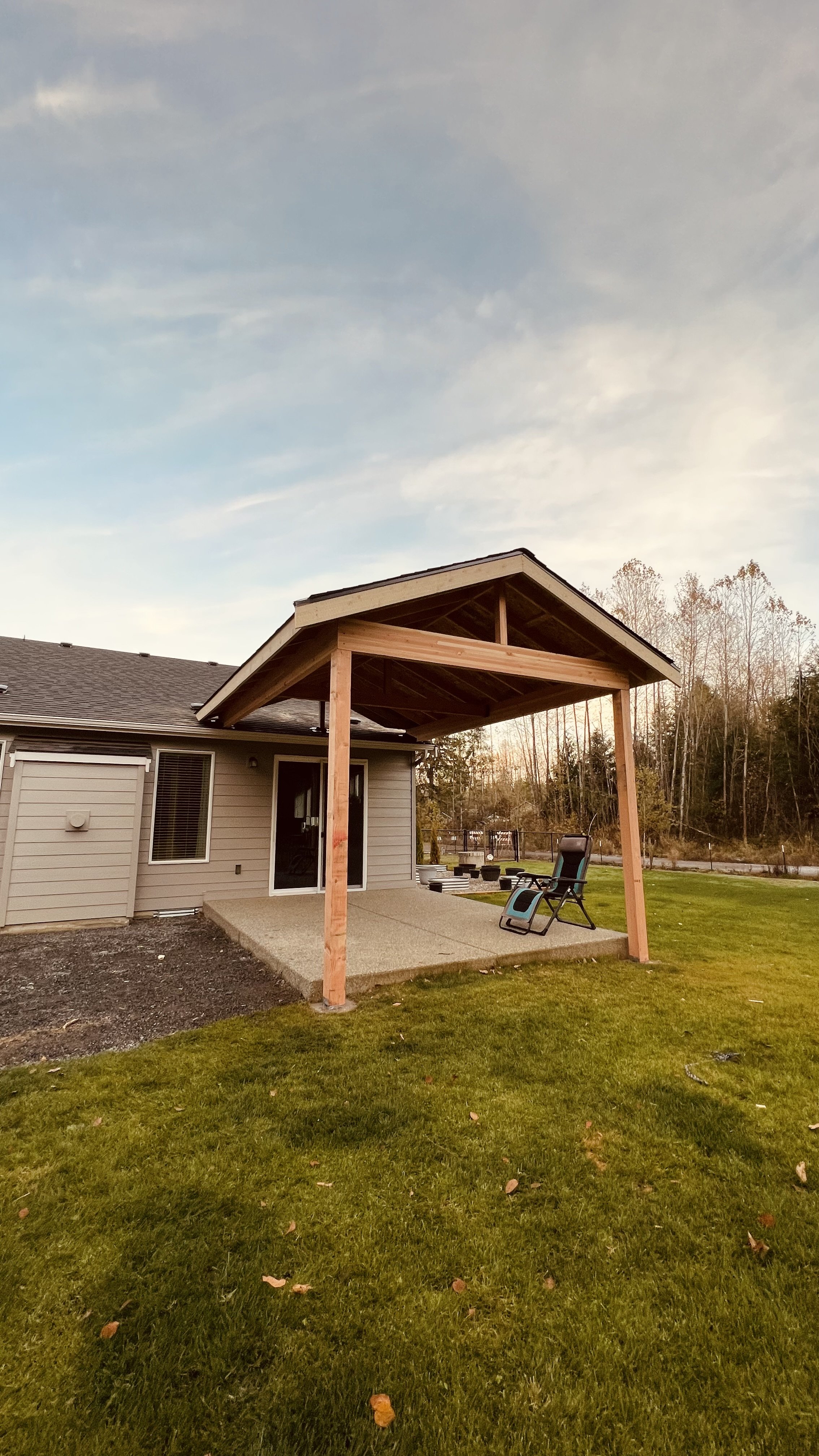 Backyard patio with a newly constructed wooden patio cover, a reclining chair, and a grassy lawn, with trees and a fence in the background during sunset.