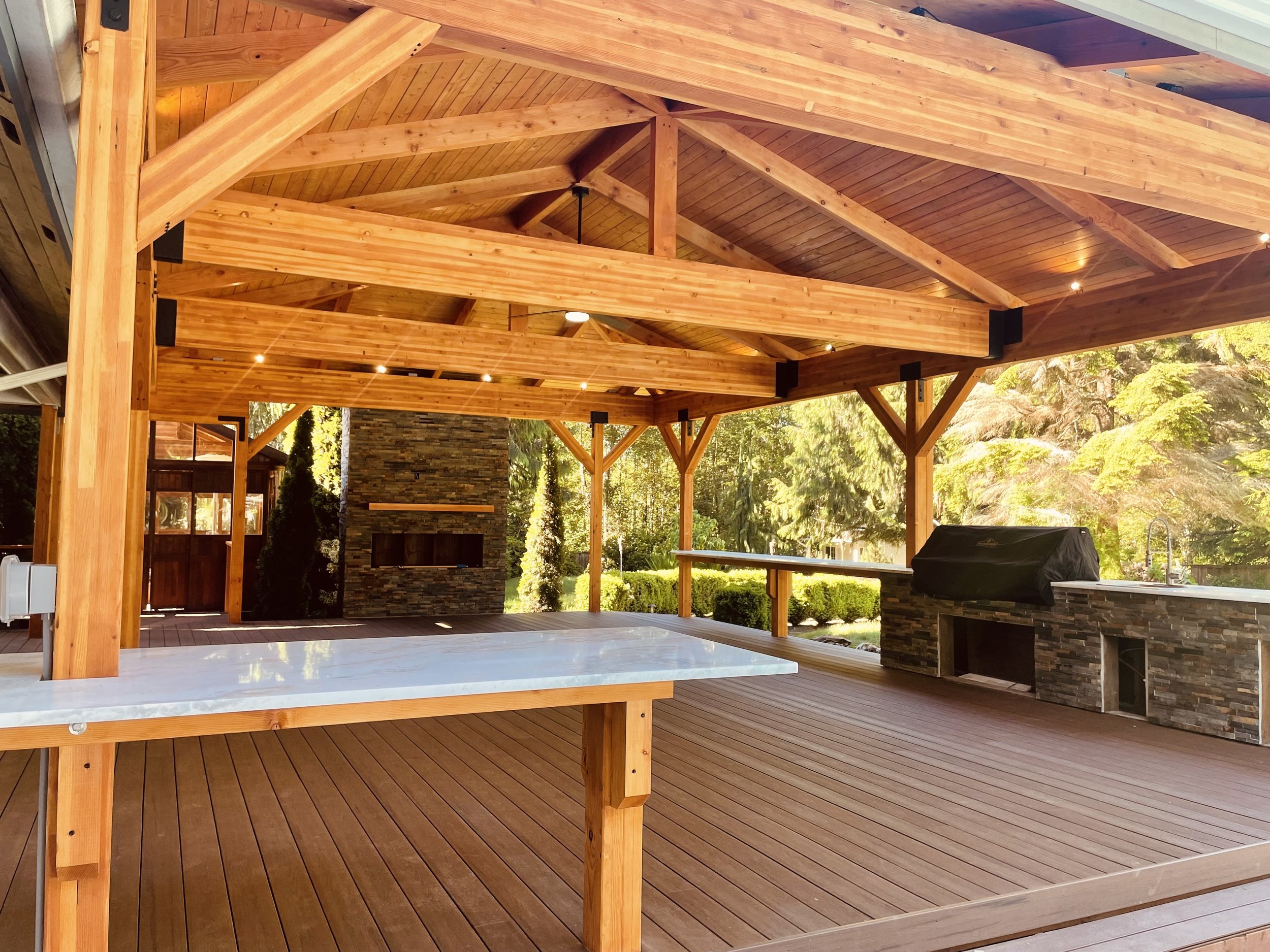 Covered outdoor patio with wooden beams, ceiling, and deck flooring, featuring a stone fireplace, sink, and barbecue grill, overlooking a lush green garden.