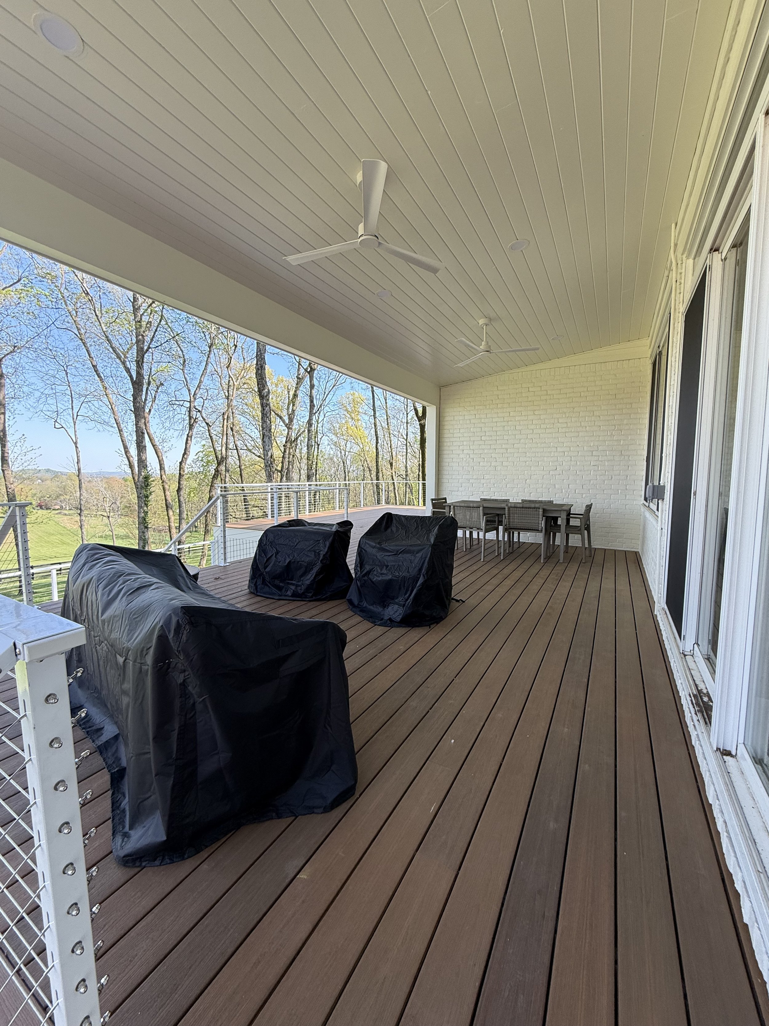 Covered outdoor deck with ceiling fans, outdoor table and chairs, and trees in the background.