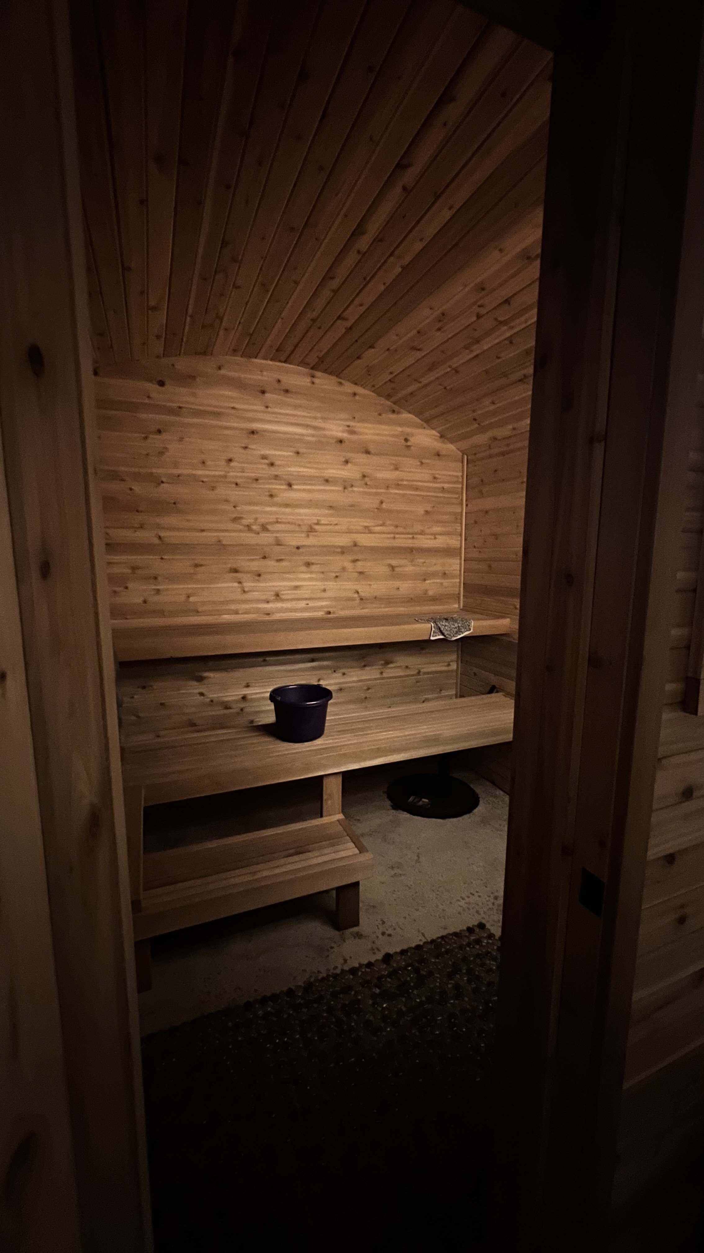 Wood-paneled interior of a sauna with a bench, a small black bucket, and a sauna heater underneath.