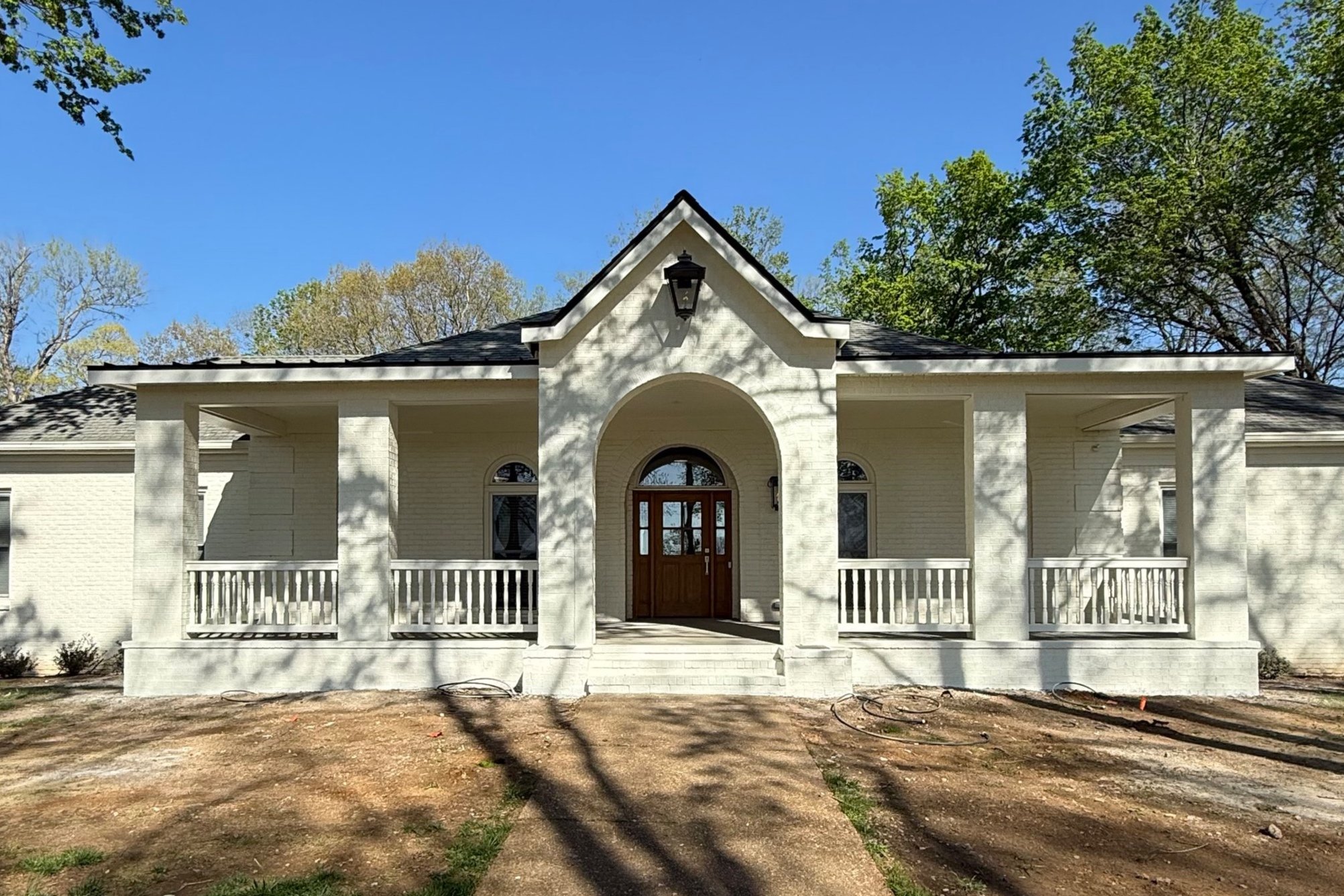 White brick house with arched entrance, front porch, and wooden front door, surrounded by trees and a clear blue sky.