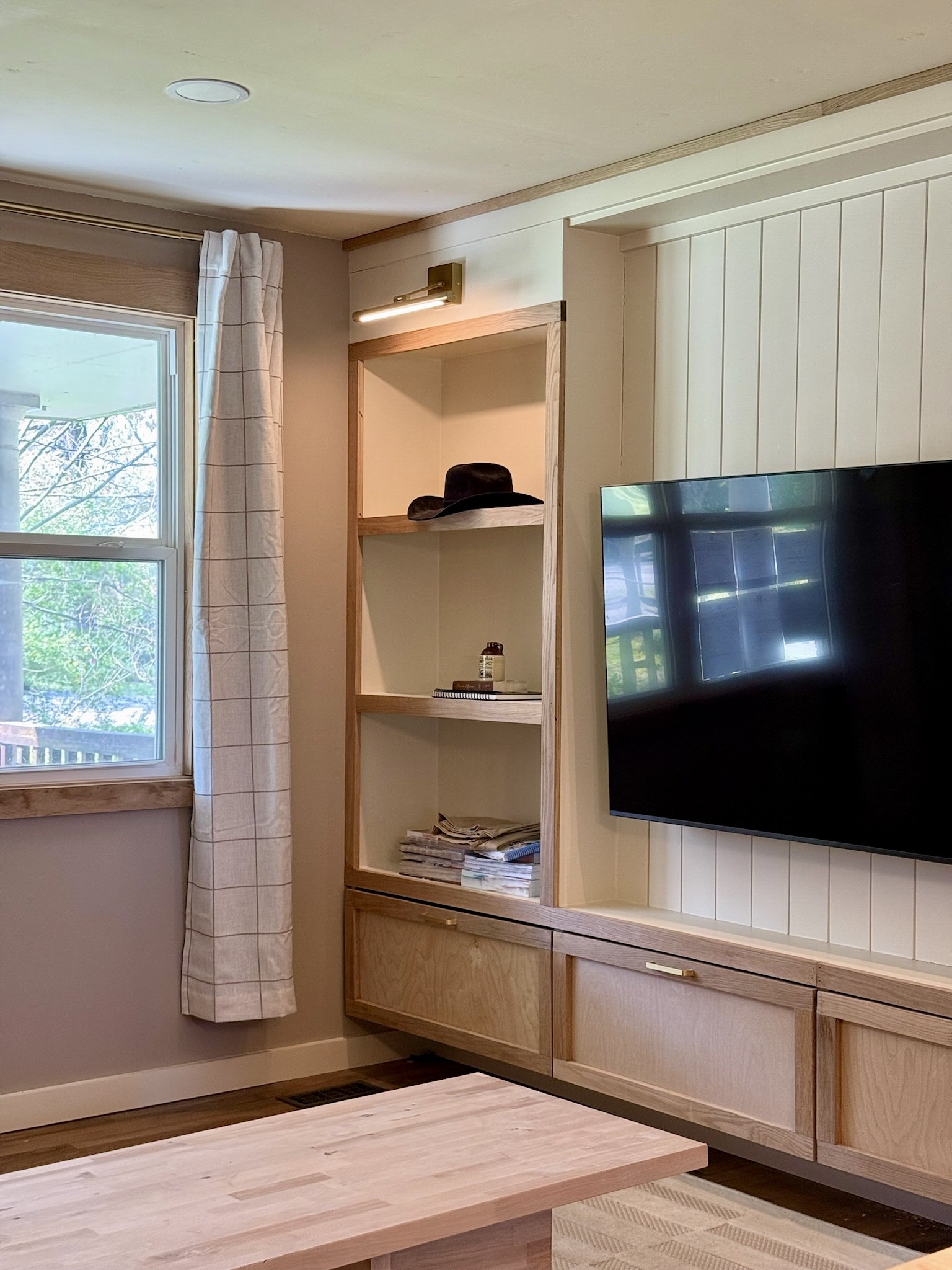Living room with a window, white curtains, wooden shelving unit containing a hat, a bottle, papers, and magazines, a wall-mounted TV, and a wooden coffee table.