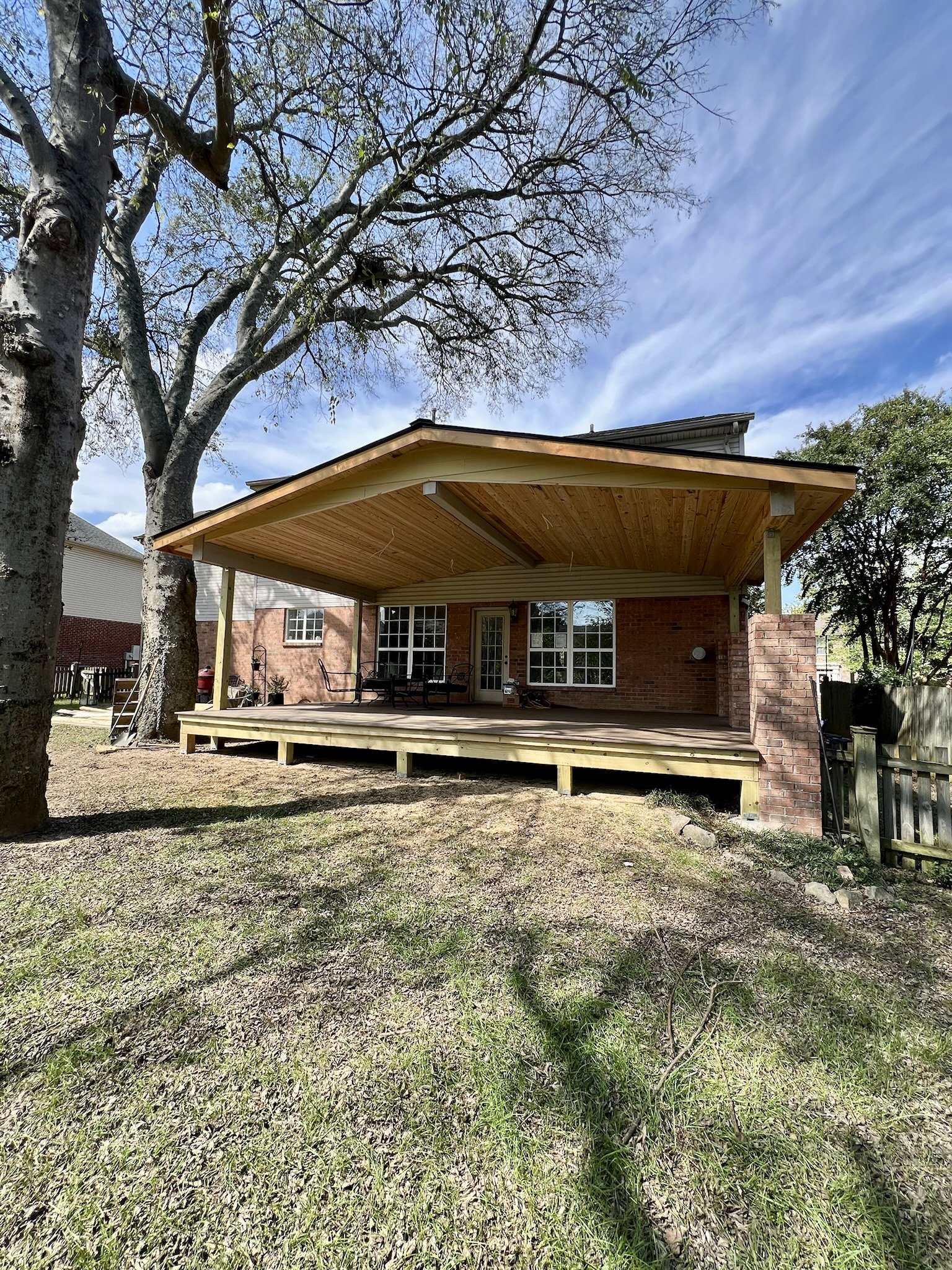 Backyard with a new wooden deck and pergola attached to a brick house, large tree, and partly cloudy sky.