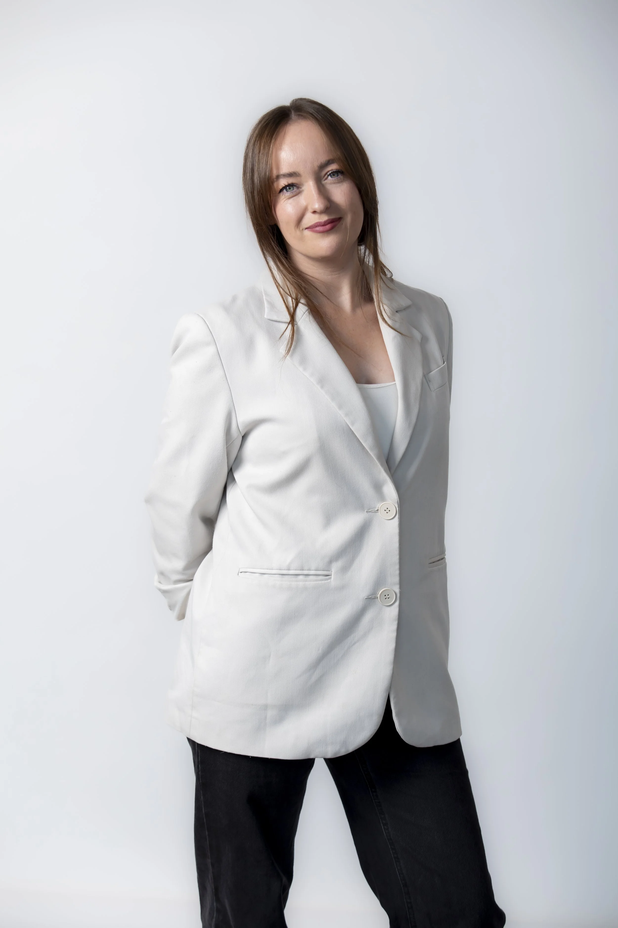 A woman with red hair, wearing a light-colored blazer and black pants, standing against a white background with a slight smile.