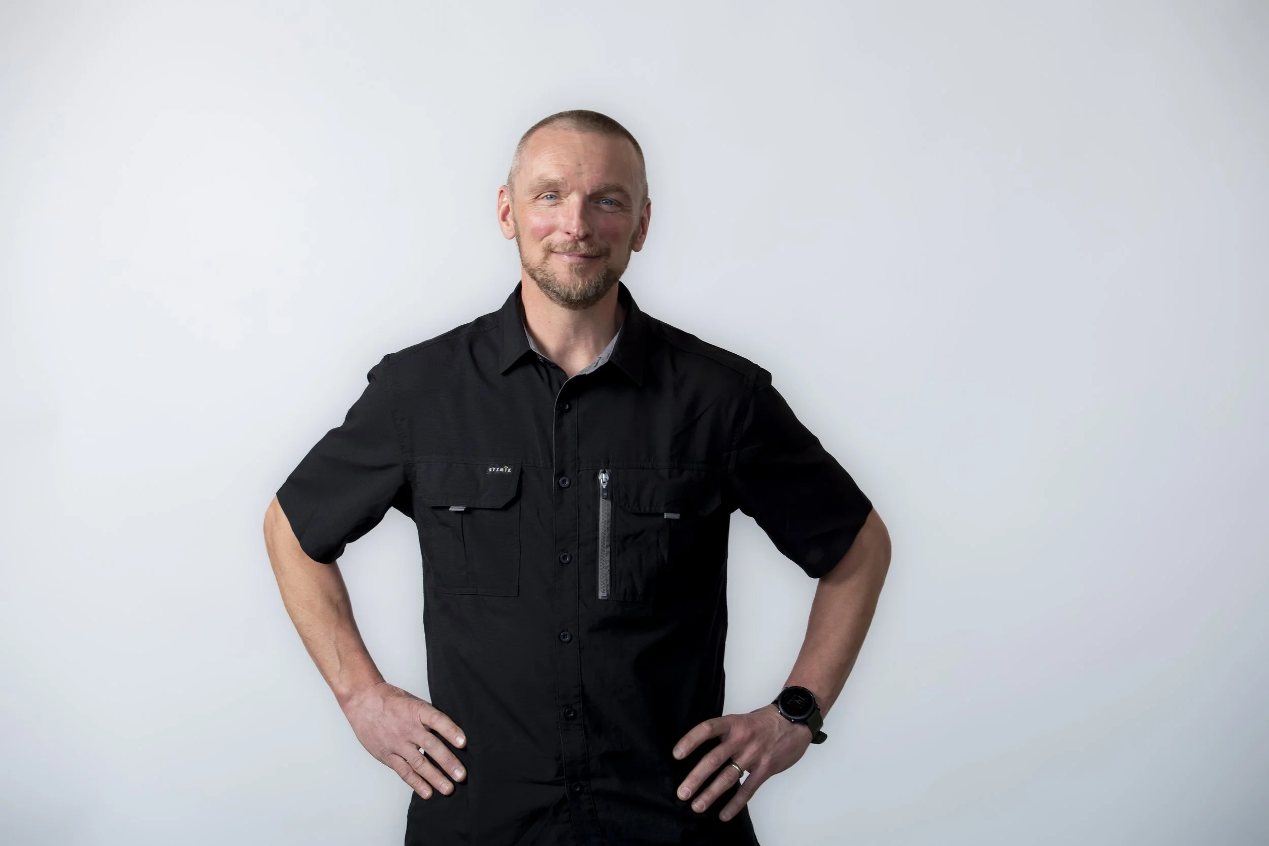 A middle-aged man with a beard and short hair, wearing a black short-sleeved button-up shirt, stands against a plain white background, smiling slightly, with hands on his hips.