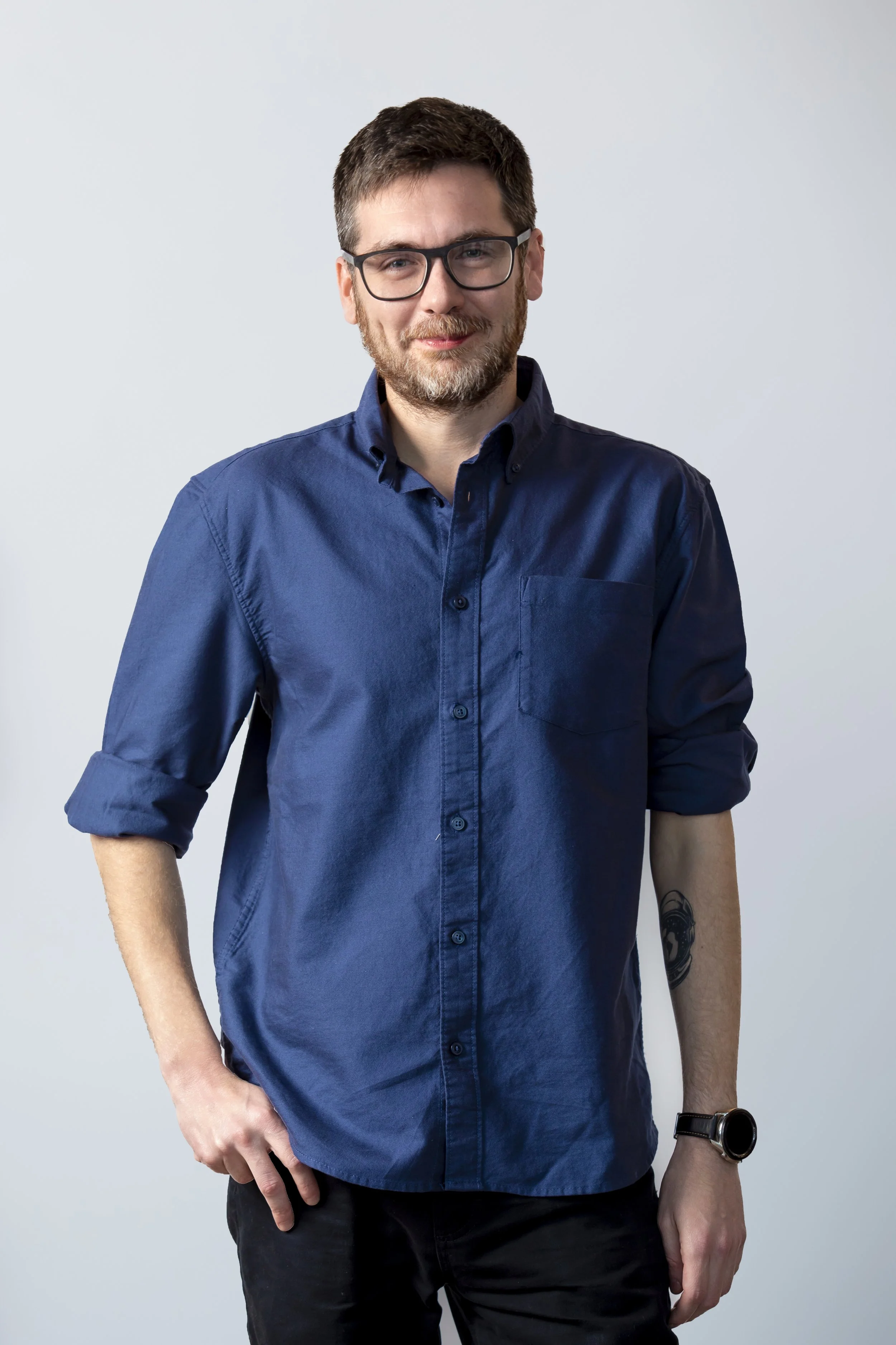 A man with glasses and a beard, wearing a blue button-up shirt with rolled sleeves, standing against a plain light-colored background.
