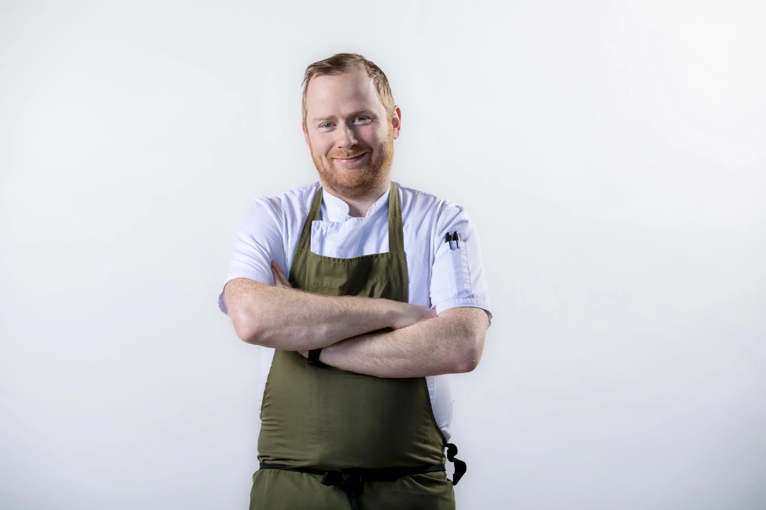 A smiling man with a beard, wearing a white chef's coat and a green apron, standing with arms crossed against a plain white background.