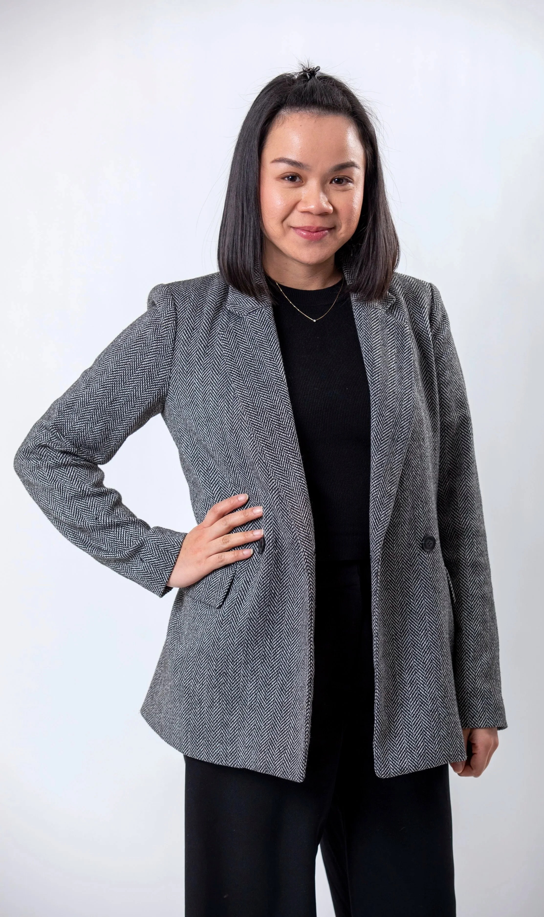 A woman with shoulder-length dark hair, wearing a gray patterned blazer over a black top, standing against a plain white background with one hand on her hip and smiling.