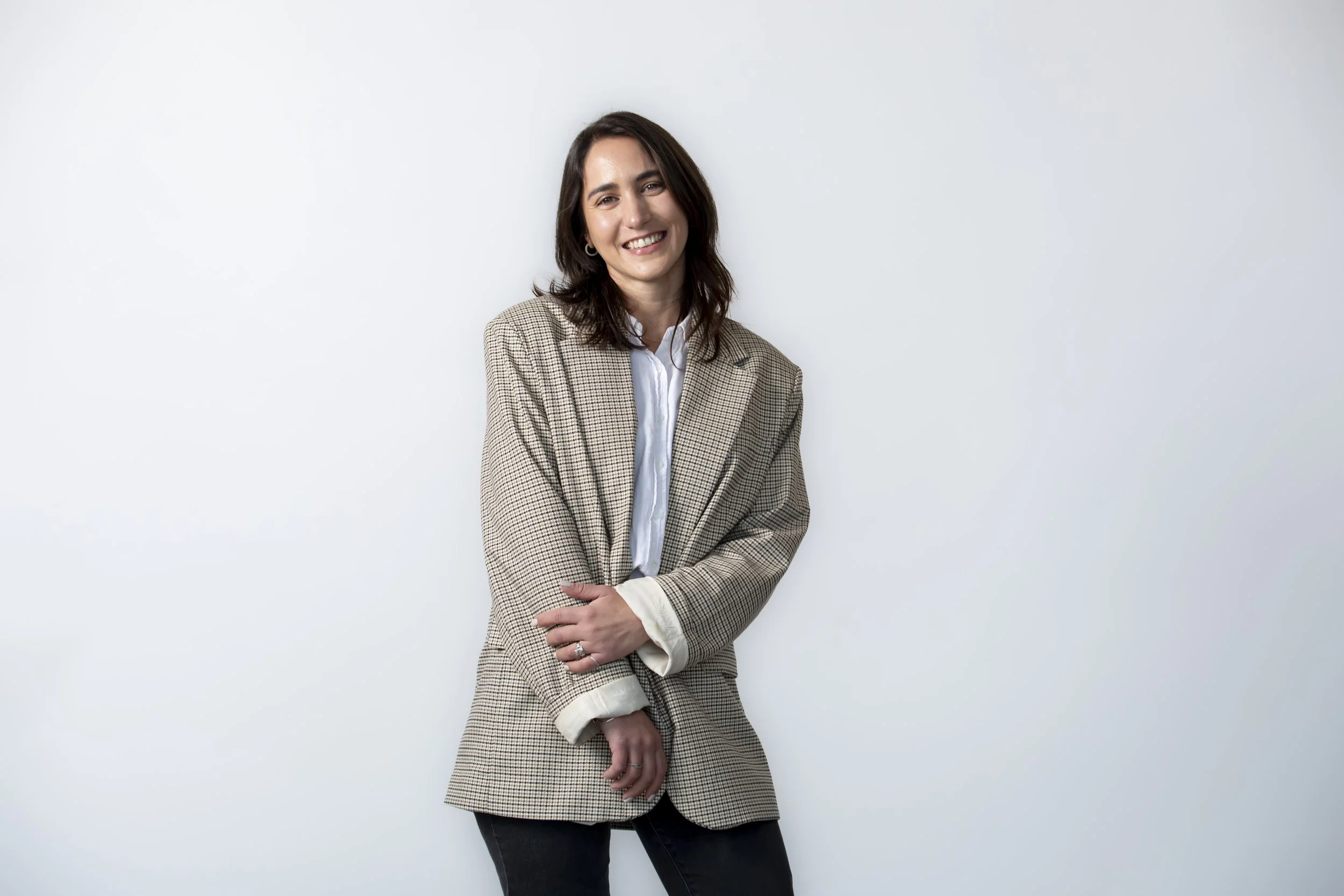 A woman with shoulder-length dark hair wearing a beige checkered blazer and a white shirt smiling and posing in front of a plain white wall.