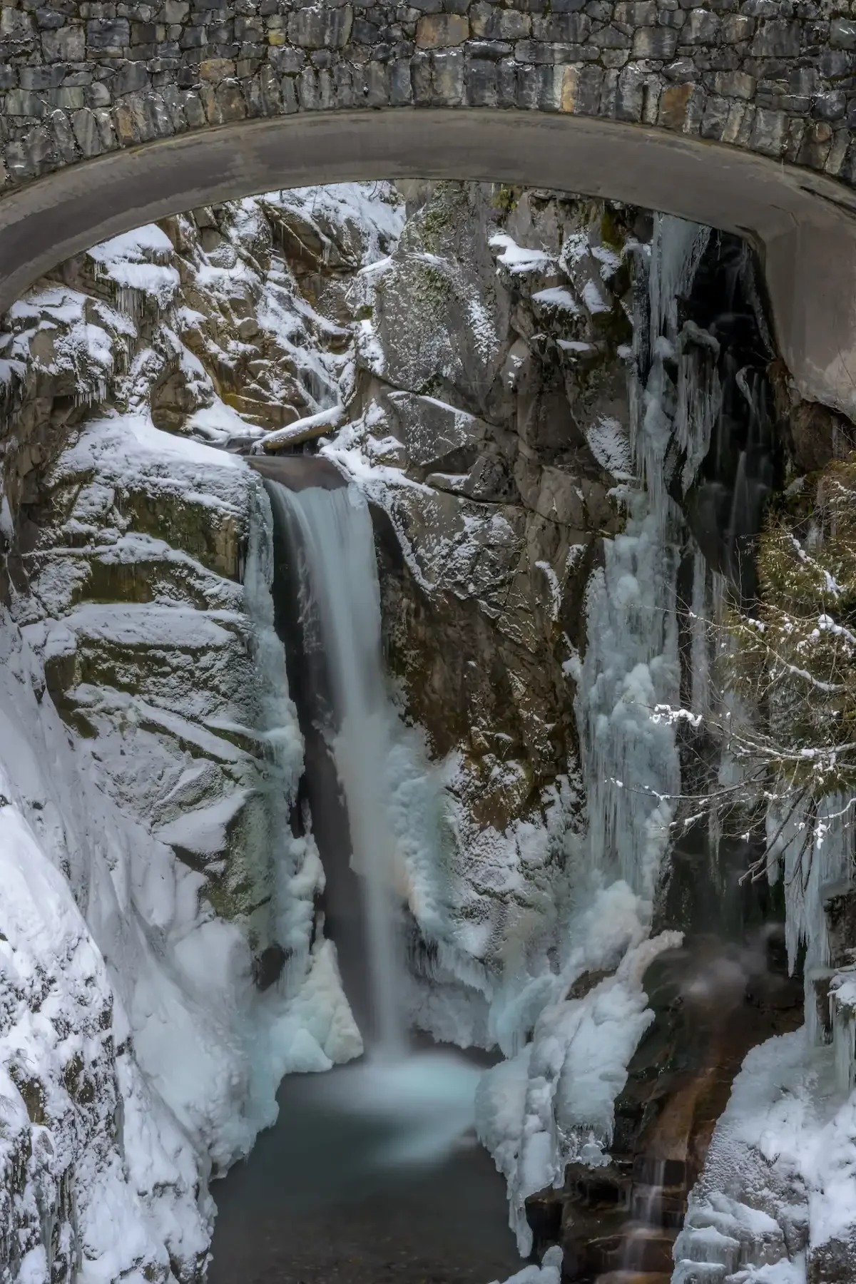Christine Falls cascading through ice and snow beneath the historic stone arch bridge at Mount Rainier National Park — winter photo stop near Fjellsangin cabin in Ashford, WA
