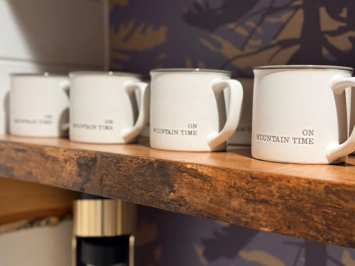 On Mountain Time ceramic mugs lined up on live-edge maple shelving at the Fjellsangin coffee bar — slow mornings at this luxury cabin near Mount Rainier in Ashford, WA