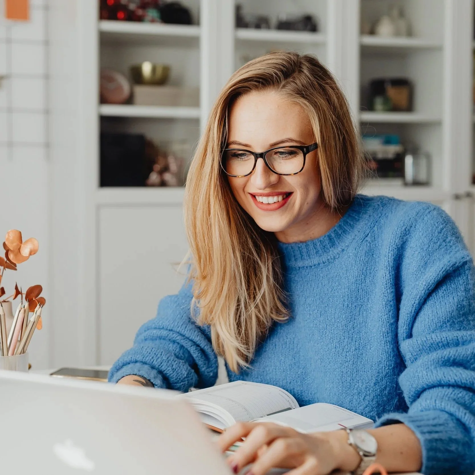 A woman with red hair, glasses, and a blue sweater is sitting at a desk, smiling and reading a book, with shelves in the background.