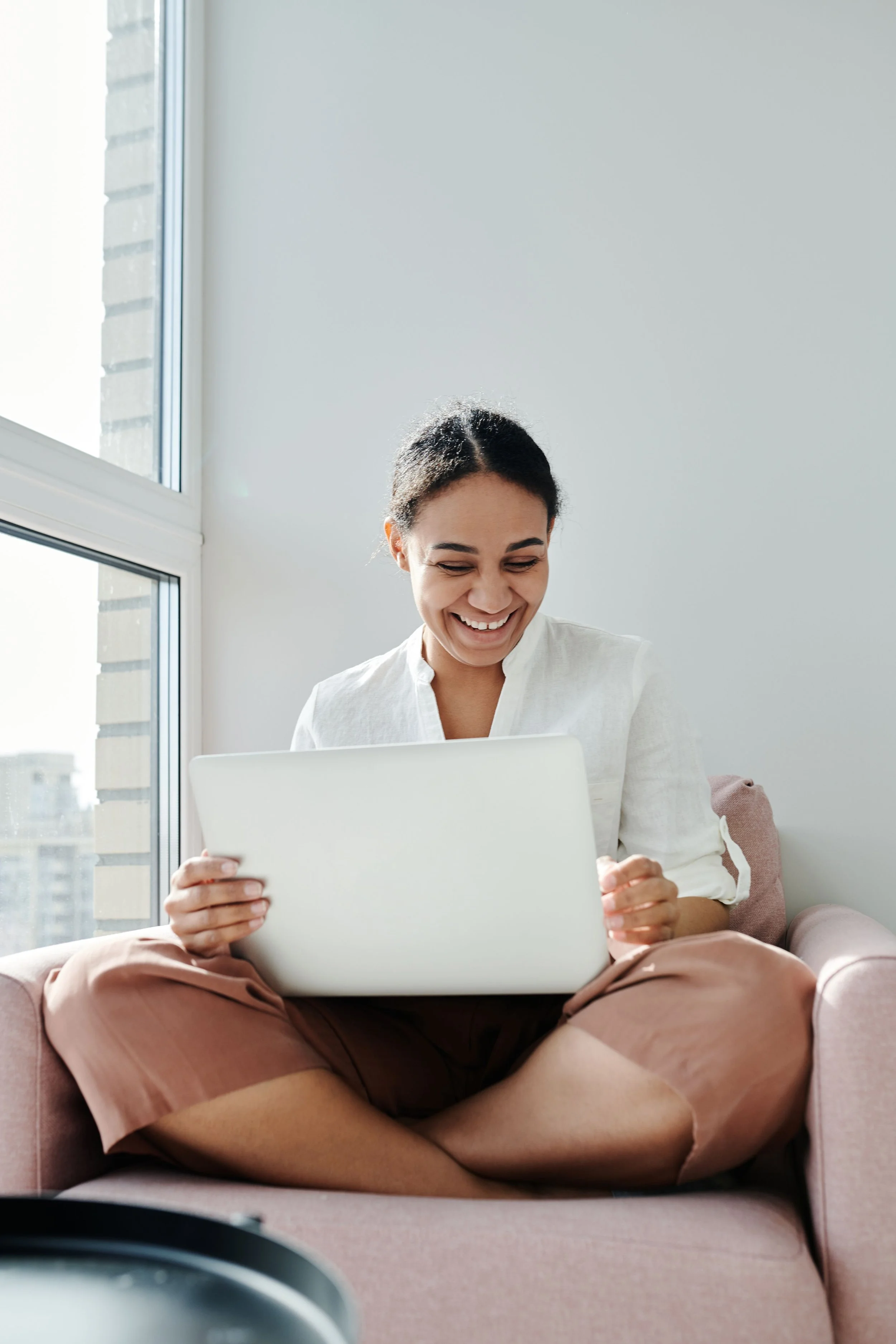 Woman smiling while sitting on a pink couch, holding a laptop, with large windows and city buildings in the background.