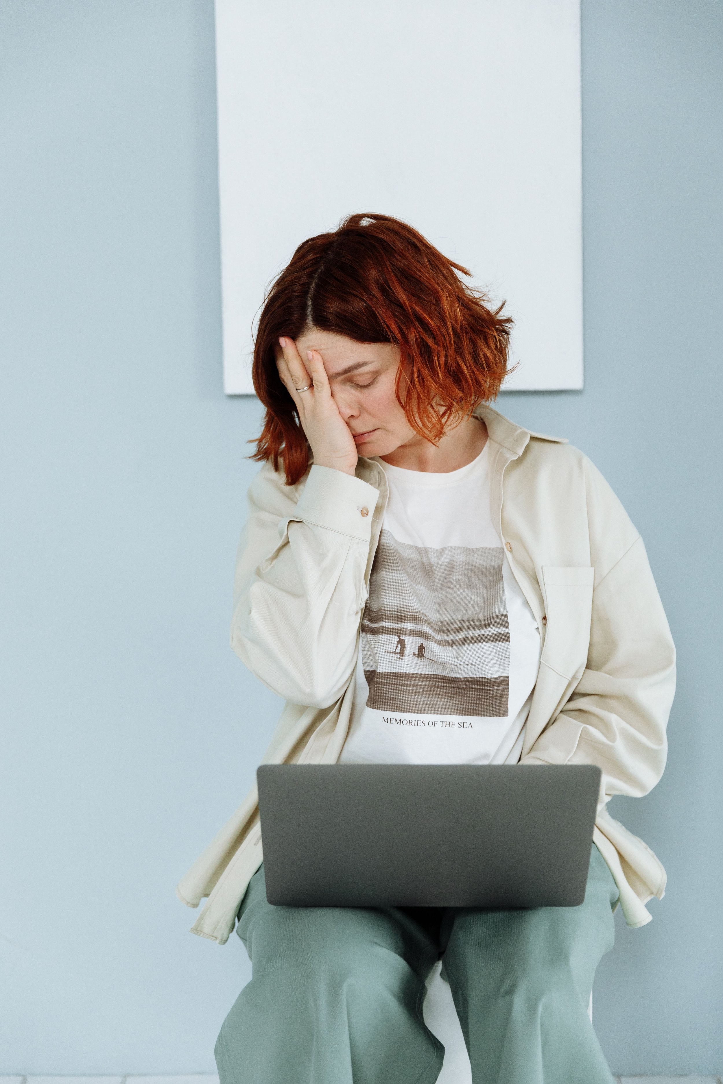 A woman sitting on a chair, holding her forehead with one hand, looking down at a laptop on her lap, with a white and gray background.
