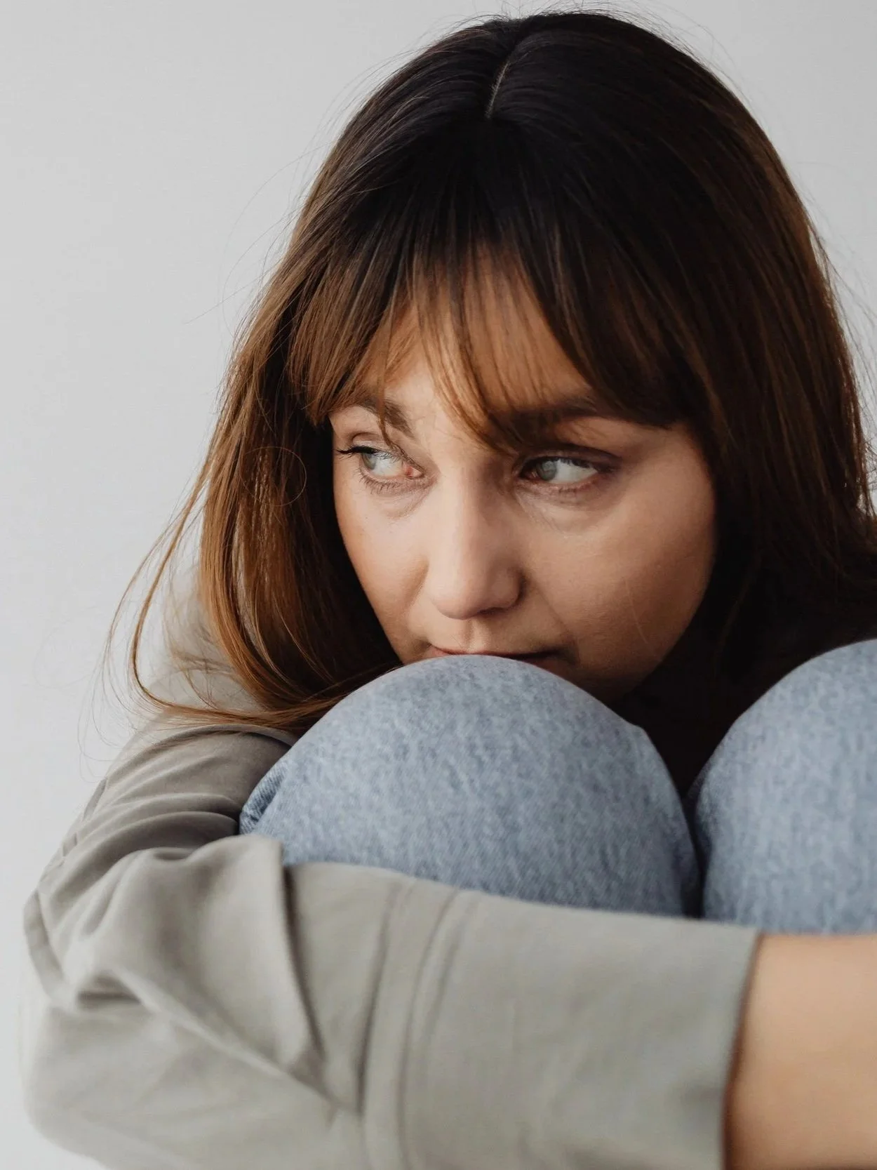 A woman with brown hair sitting in a contemplative pose, wearing a beige jacket and gray pants, with her knees pulled close to her chest against a plain light background.