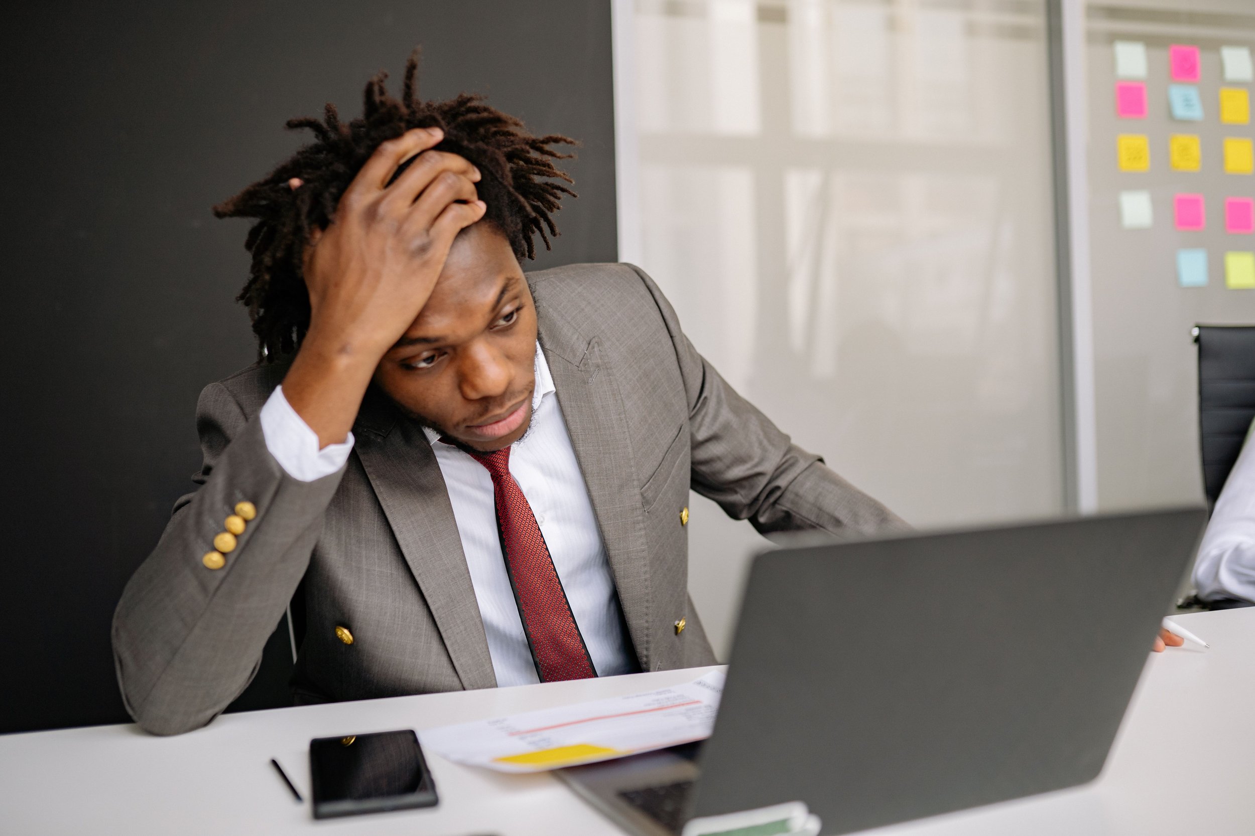 A man in a gray business suit and red tie sitting at a desk with his hand on his forehead, looking worried or stressed, in an office setting.