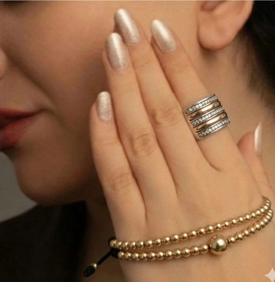 Mujer con uñas pintadas de dorado, mostrando varios anillos en los dedos y una pulsera de perlas en la muñeca.