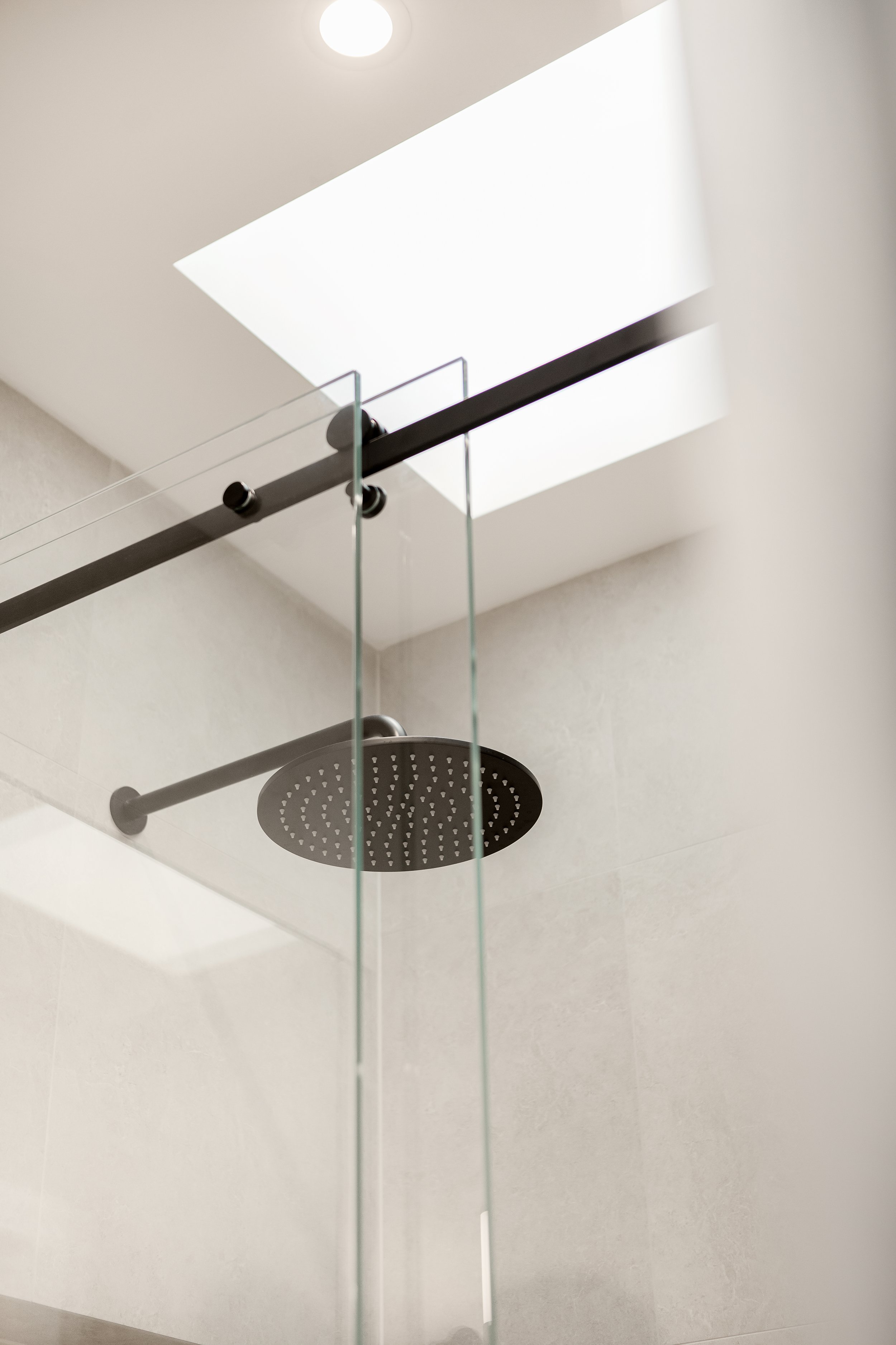 Luxury residential bathroom renovation detail featuring frameless glass sliding shower doors with matte black hardware, a round black rainfall shower head, and neutral grey wall tiles illuminated by a skylight.