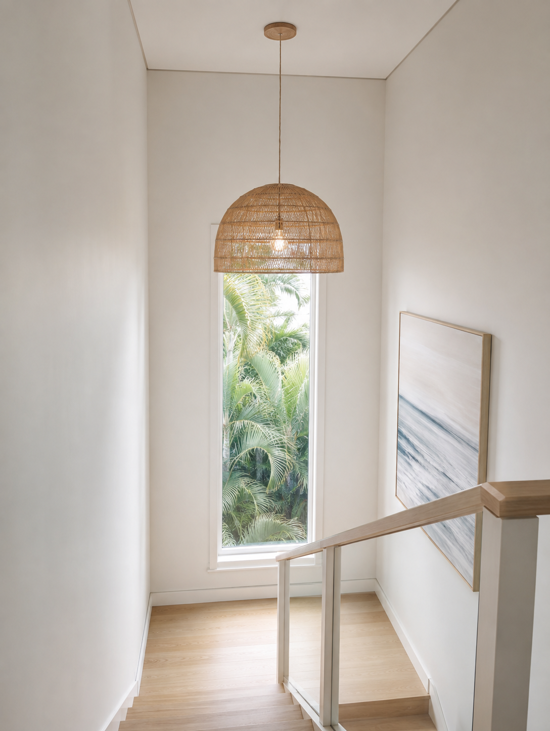 Modern coastal home stairwell design featuring a large woven rattan dome pendant light, light oak timber stairs, a vertical window framing palm trees, and ocean wave artwork.