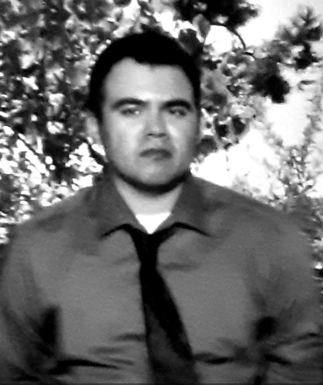 Black and white photo of a man with short dark hair, wearing a collared shirt and a tie, standing outdoors with trees in the background.