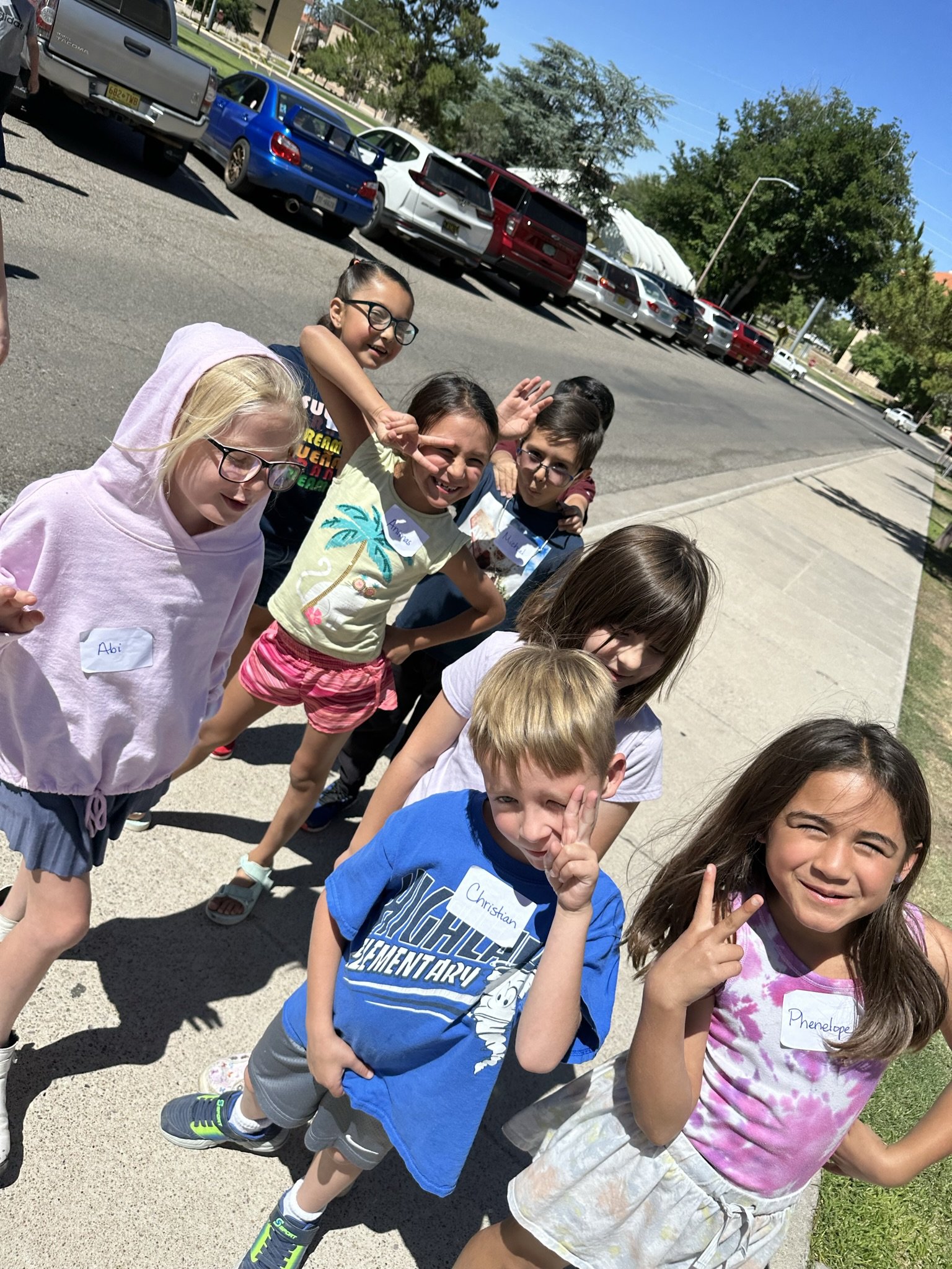 Group of children standing on sidewalk, wearing casual clothes, some with name tags, smiling and making peace signs, sunny day with parked cars and trees in background.
