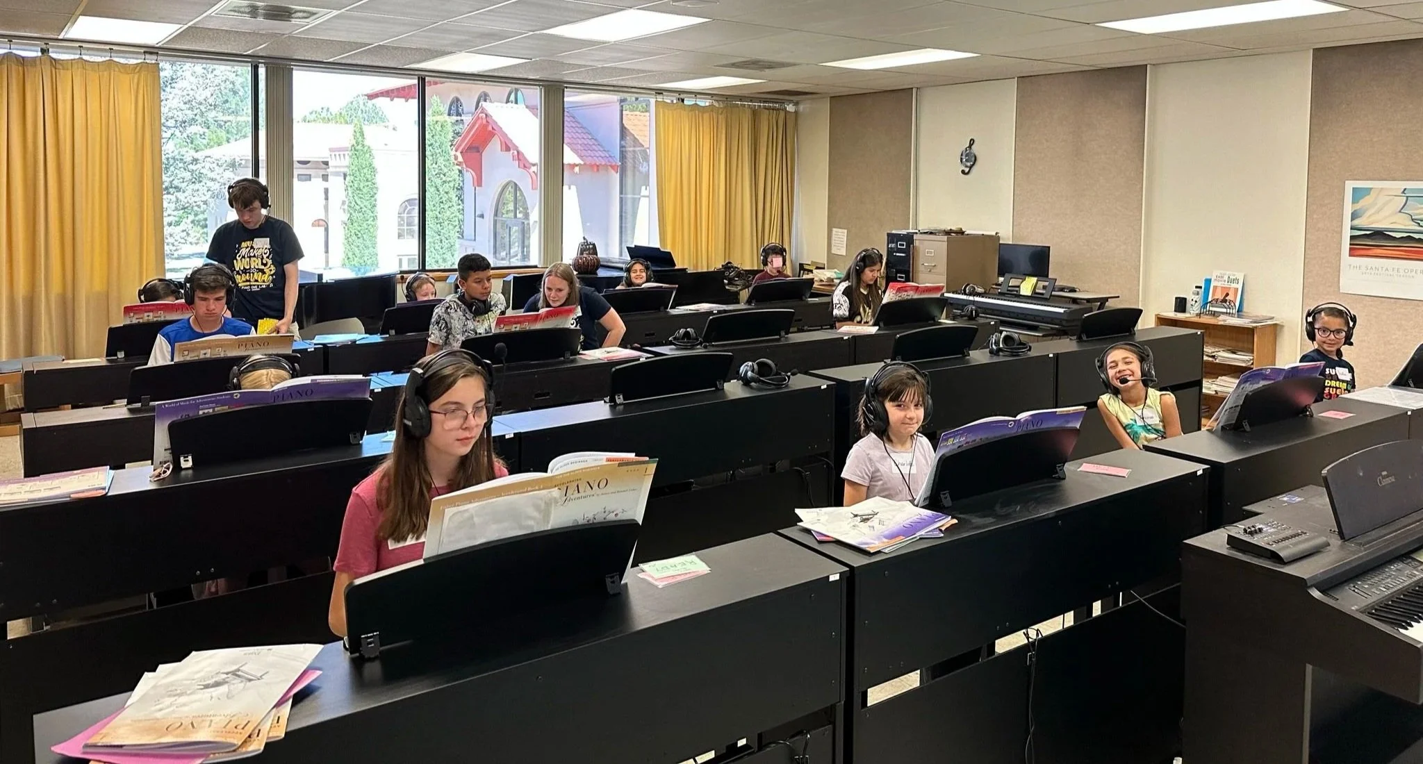 Children in a music classroom with pianos, wearing headphones, seated at their desks, with a teacher assisting one student, large windows showing trees outside, yellow curtains, and music sheets on desks.