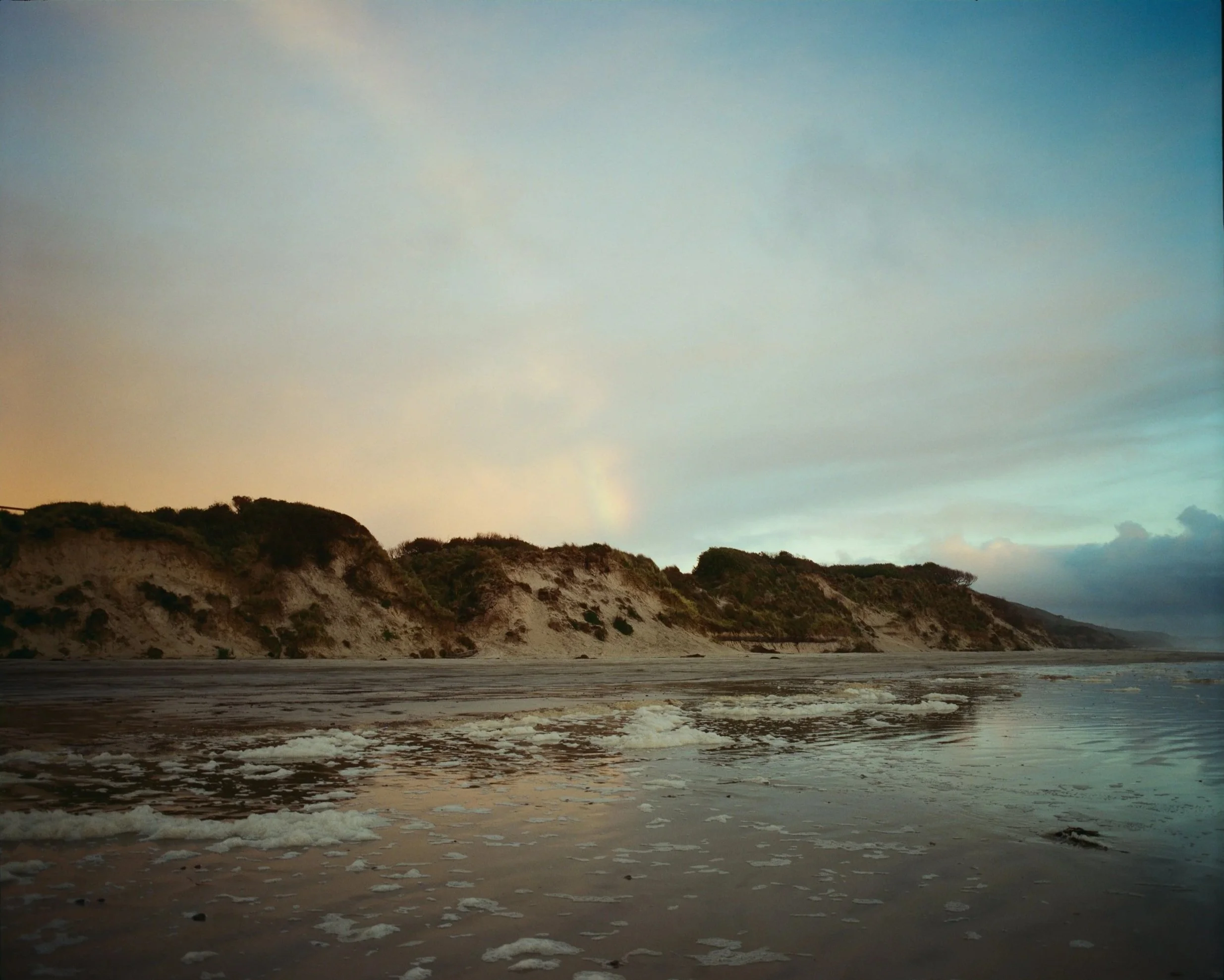 A beach with waves washing up on the shore, with sandy cliffs and vegetation in the background under a blue sky with some clouds.