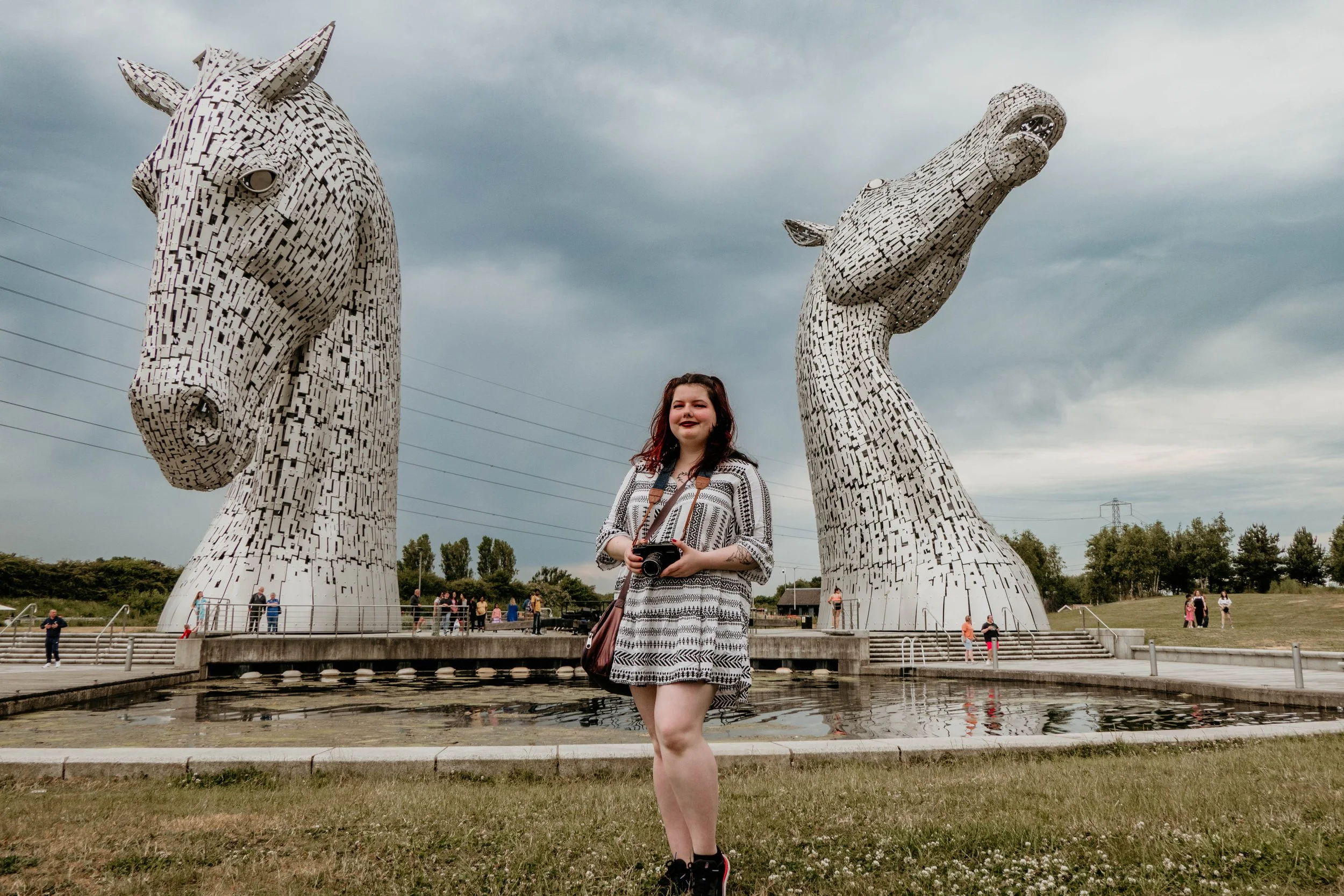 Woman standing in front of large horse head sculptures, known as The Kelpies, with overcast sky.