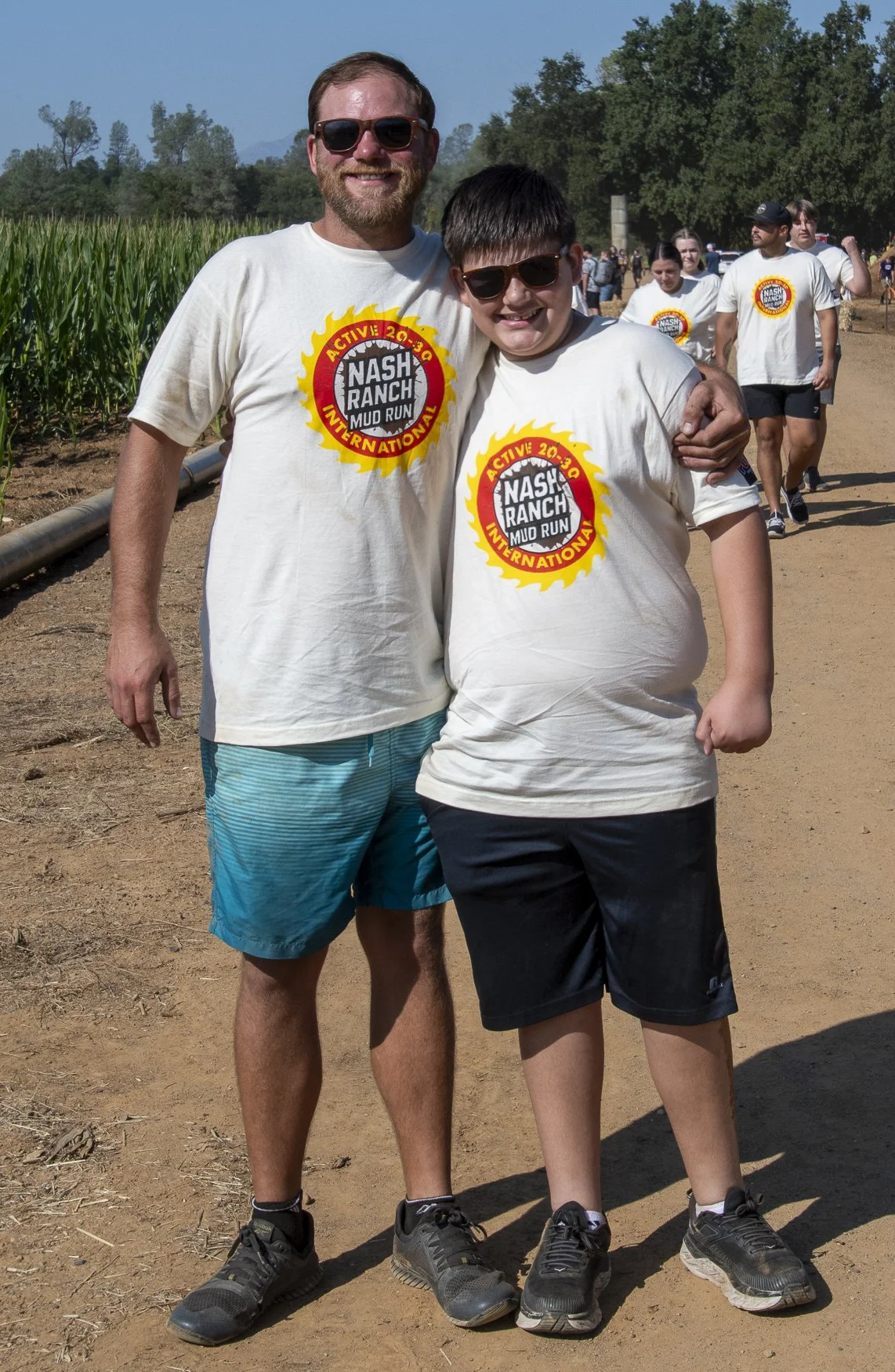 Two smiling boys wearing matching white T-shirts with a yellow, red, and black logo of the Nash Ranch Mud Run, standing outdoors on a dirt path with more participants in the background.