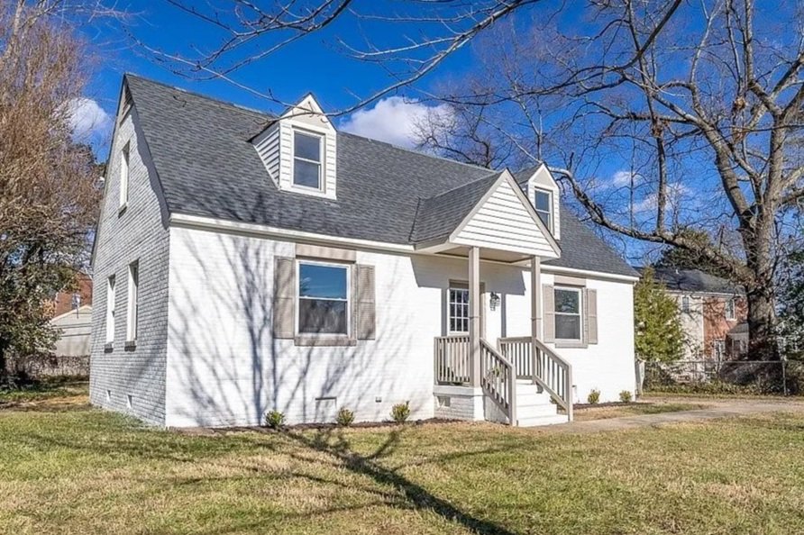 A white, two-story house with a gable roof and small front porch, surrounded by a lawn with some small plants. The house has two dormer windows on the roof and is set against a blue sky with some clouds, with leafless trees in the yard.