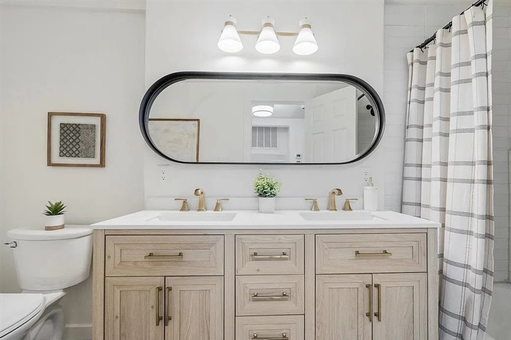 Bathroom with a double vanity, large oval mirror, three white light fixtures, white countertop, two gold faucets, a potted plant, framed artwork, a toilet with a small plant on top, and a shower curtain with a gray checked pattern.