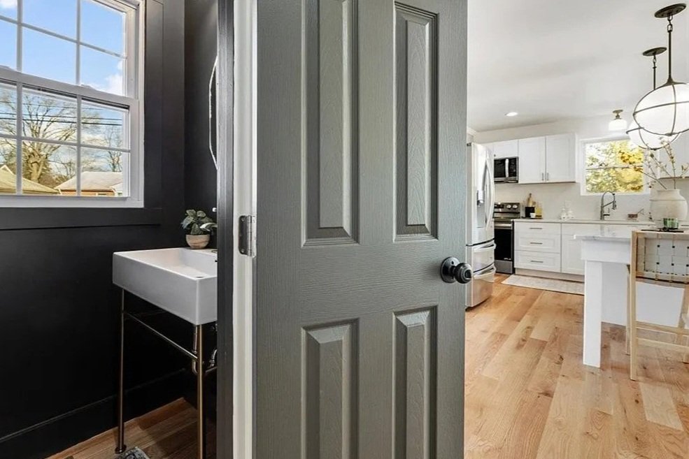 View of a modern kitchen with white cabinets, stainless steel appliances, and a wooden floor, seen through a partially open gray door. A small washbasin with a plant is visible on the left side, near a window.