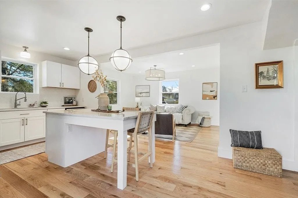 Bright and airy open-concept kitchen and living area with white cabinetry, pendant lighting, wooden flooring, and minimal decor.