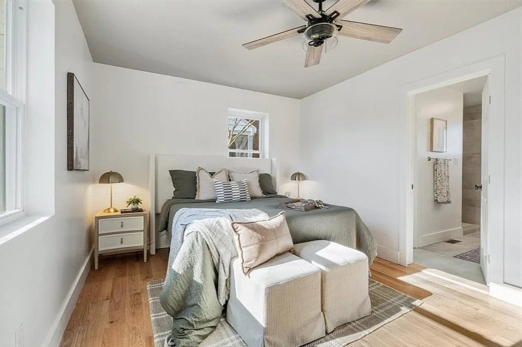 A cozy bedroom with a bed, side tables, lamps, and a ceiling fan, with a doorway to a bathroom and a window overlooking trees.