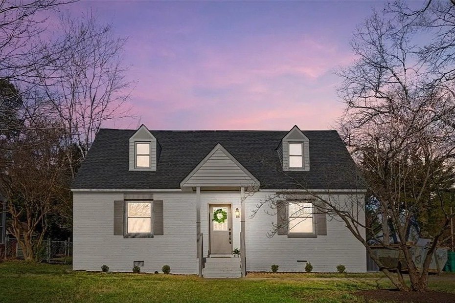 White house with gray shutters, a central front door adorned with a wreath, black shingle roof, two small dormer windows, and a front yard with grass and leafless trees at sunset.