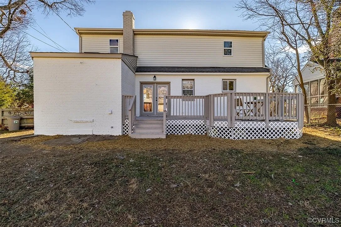 Rear view of a two-story house with a wooden deck and outdoor seating area, surrounded by trees and a wire fence.