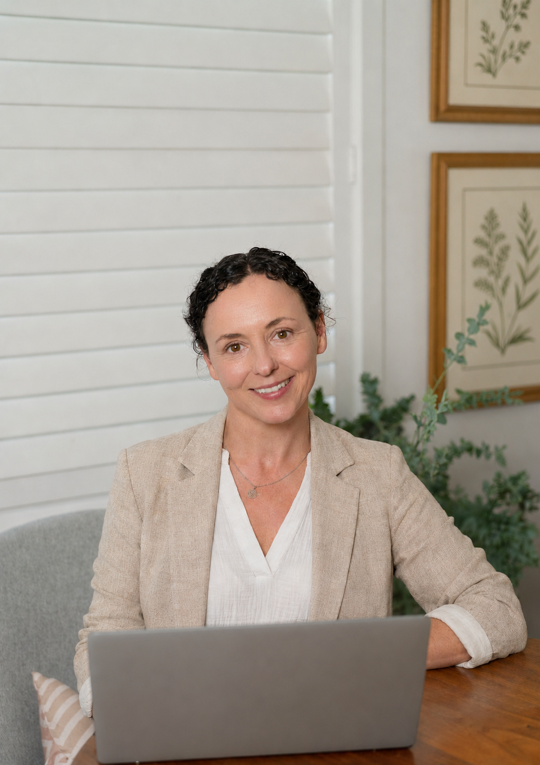 A woman with dark curly hair smiling at the camera, sitting at a desk with a laptop, in a room with light colored walls, framed botanical prints, and a green plant in the background.