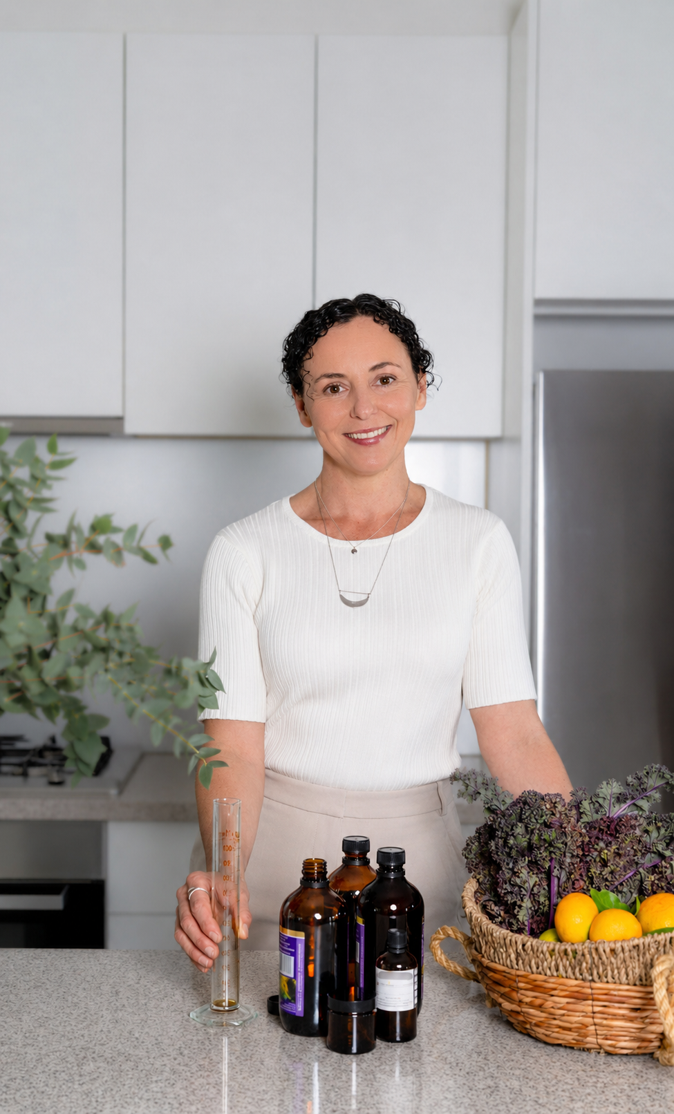 A woman with curly hair smiling in a modern kitchen, surrounded by bottles of herbal medicine, fresh vegetables, and a measuring beaker on the counter.