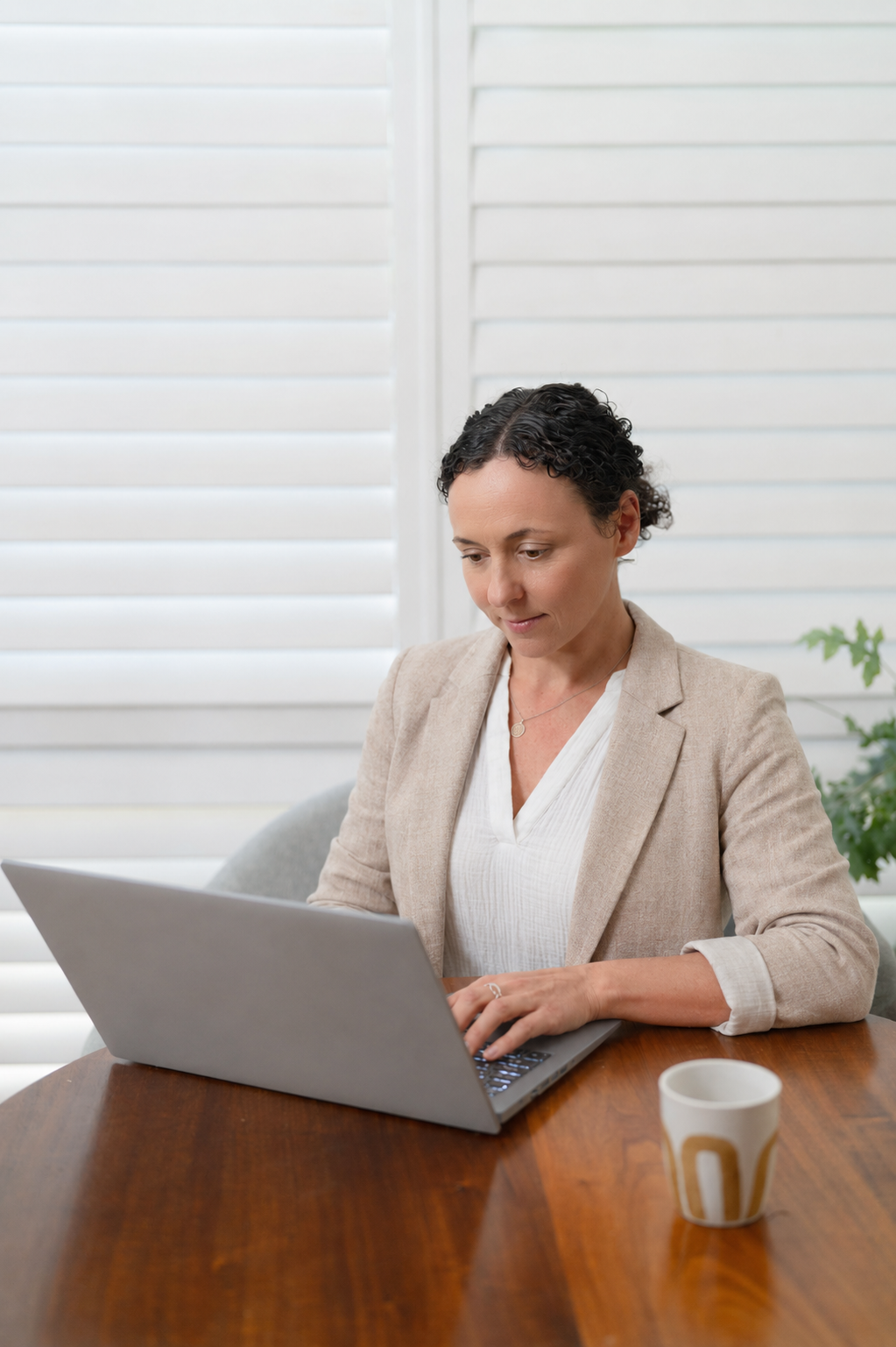A woman with short curly dark hair wearing a beige blazer and white blouse working on a silver laptop at a wooden table with a white and gold mug nearby. Background includes white window shutters and a green plant.