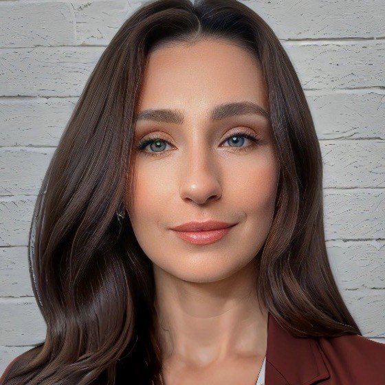 Close-up photo of a woman with long brown hair, blue eyes, and a subtle smile, standing in front of a white brick wall.