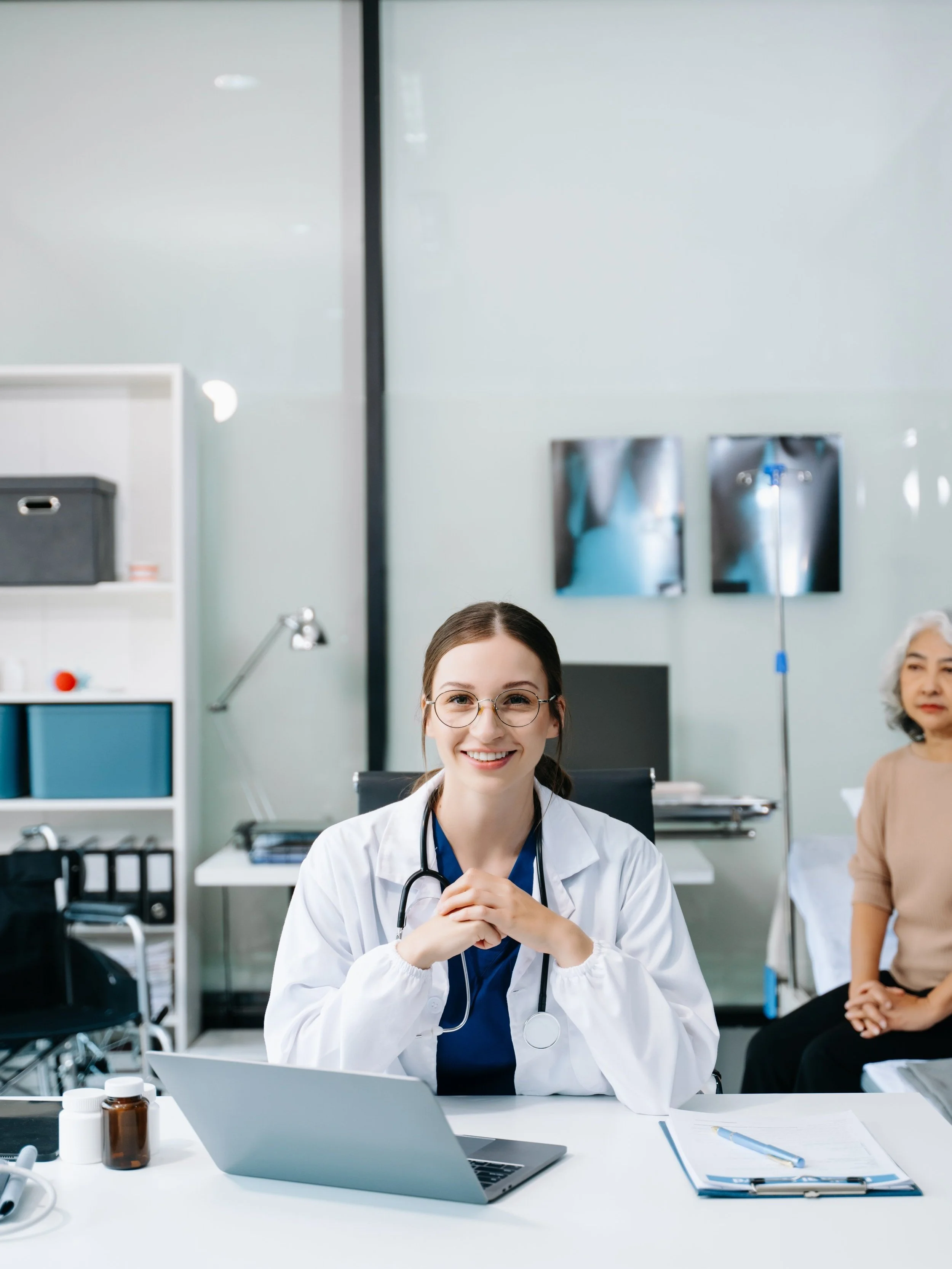 Doctor smiling at desk in medical office with patient sitting in the background, stethoscope around neck, laptop and clipboard on desk.