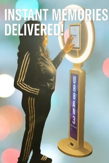 A woman in a black jacket and striped pants using a touchscreen kiosk with the sign 'Urban 3' and the text 'Instant Memories Delivered!' above.