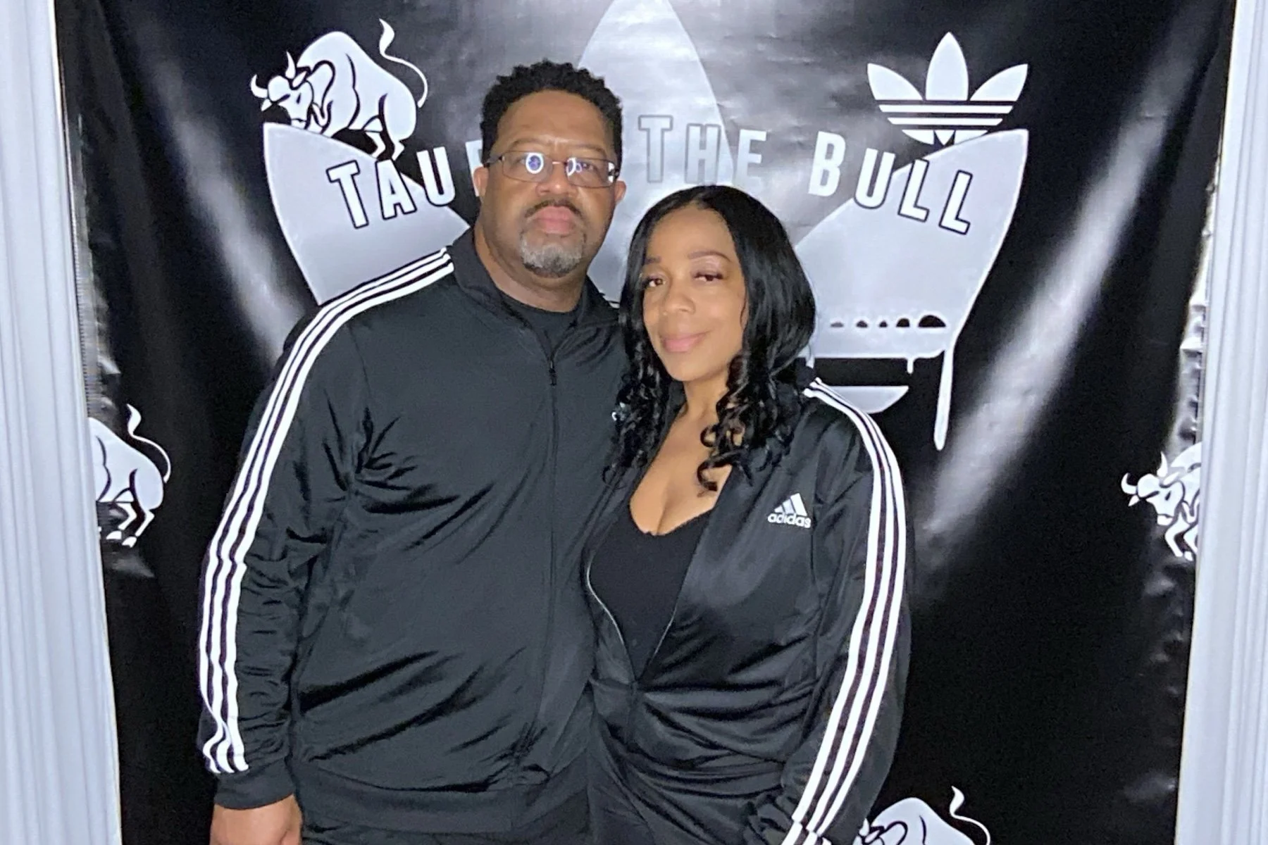 A man and woman wearing black Adidas jackets with white stripes on the sleeves, standing in front of a black backdrop featuring a stylized bull's head, the words "TAUR THE BULL," and Adidas logo.