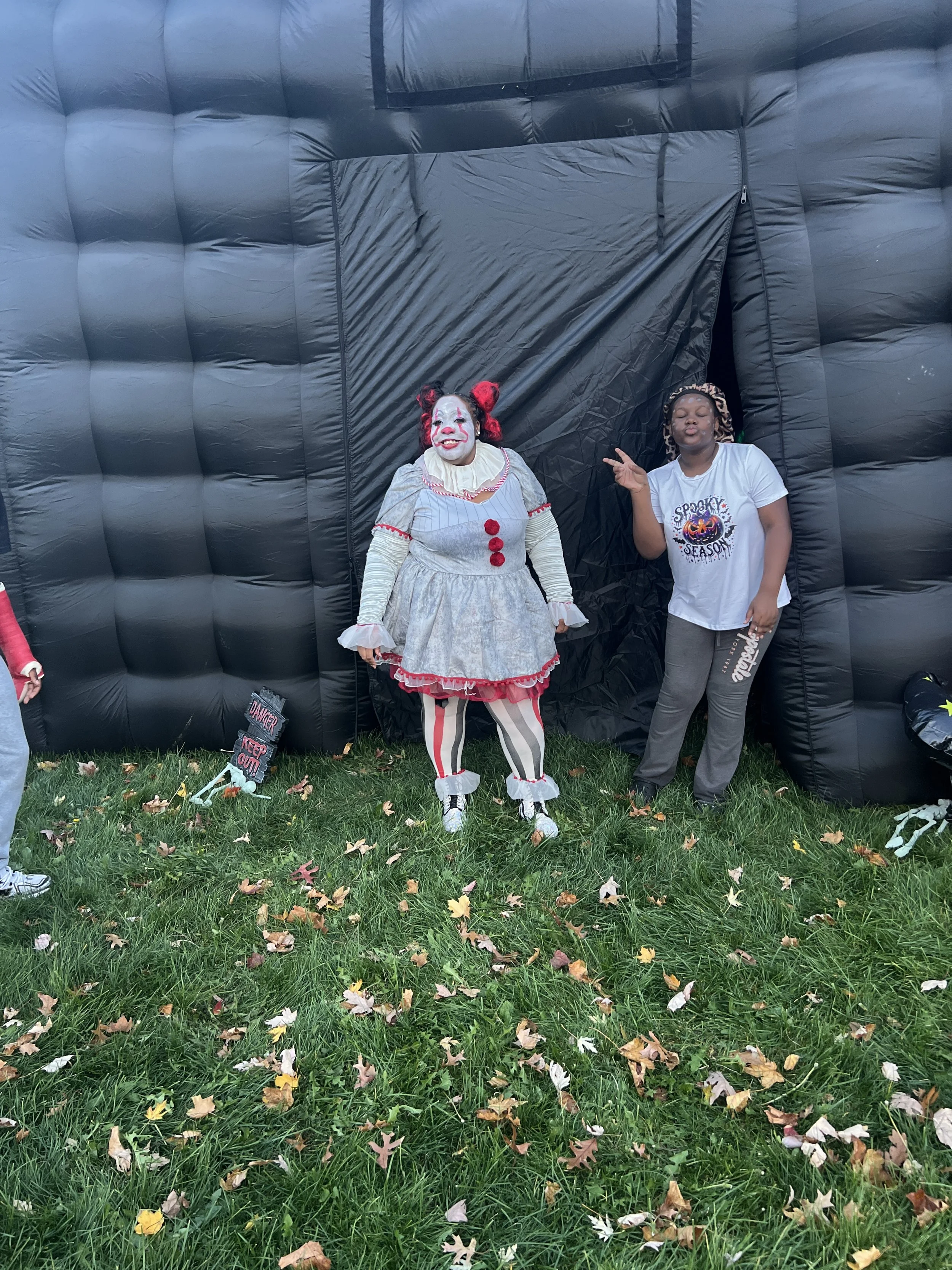 A person dressed as a scary clown and a girl posing next to a black inflatable haunted house outdoor during Halloween.