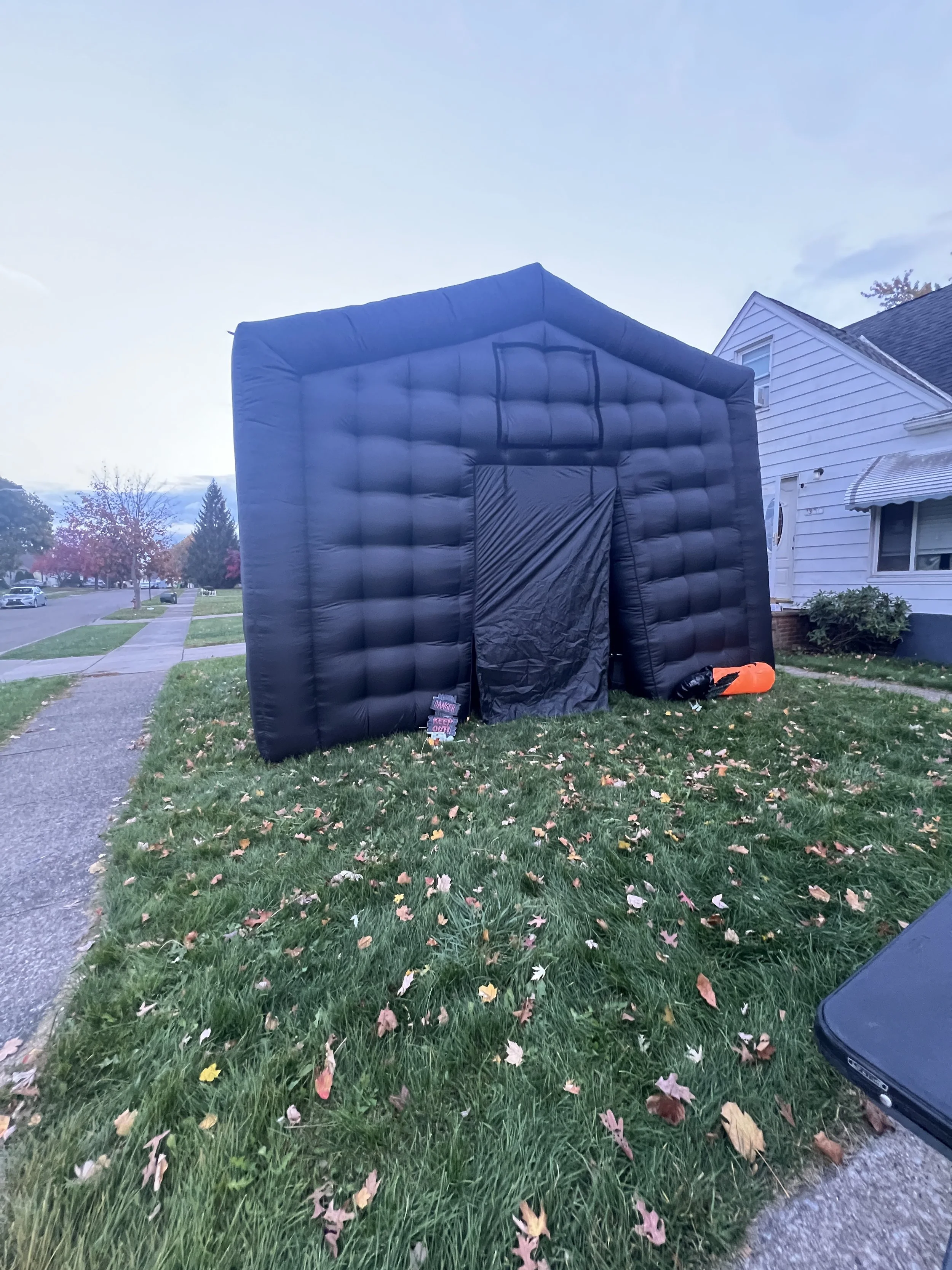 Inflatable black house structure on a lawn with fallen leaves, houses, and a sidewalk in the background.