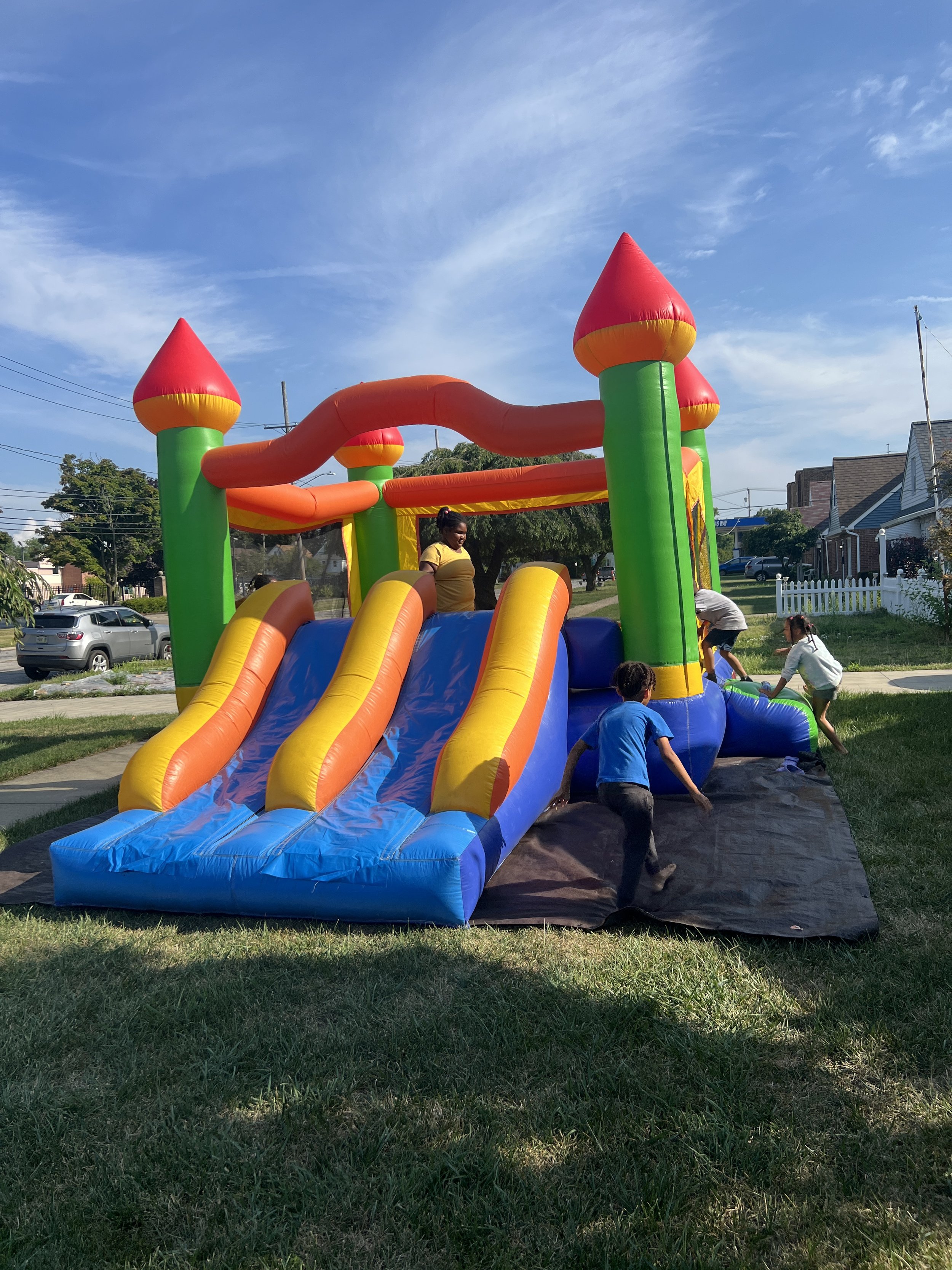 Children playing on a colorful inflatable bounce house with slides, shaped like a castle with red turrets and orange towers, set up on a grassy yard on a sunny day.