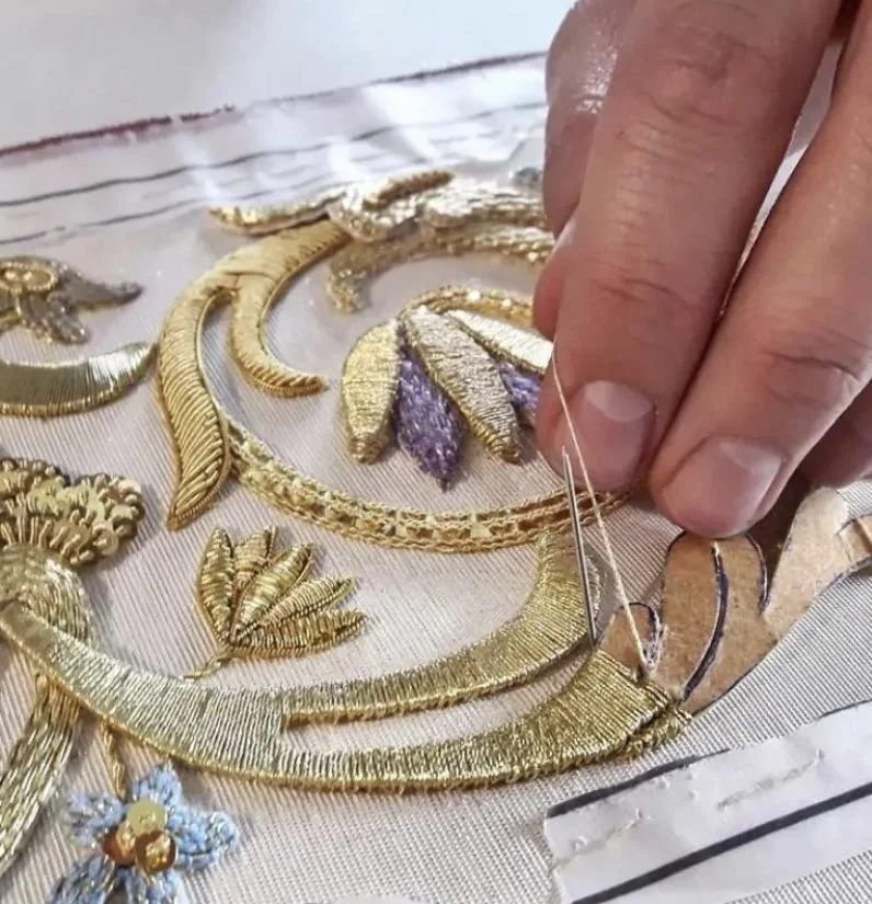 A person sewing intricate gold embroidery onto fabric, creating a floral pattern with details in purple and gold.