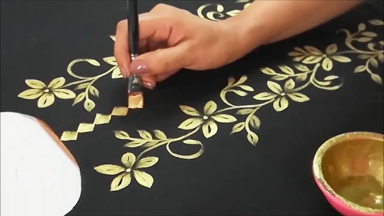 Close-up of a hand painting a floral design with gold paint on a black surface, with a small bowl of gold paint nearby.