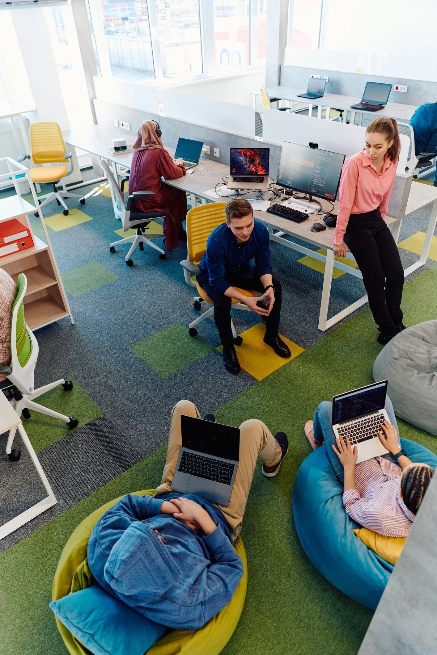 People working and relaxing in a modern office space with colorful carpet, bean bags, desks, and large windows.